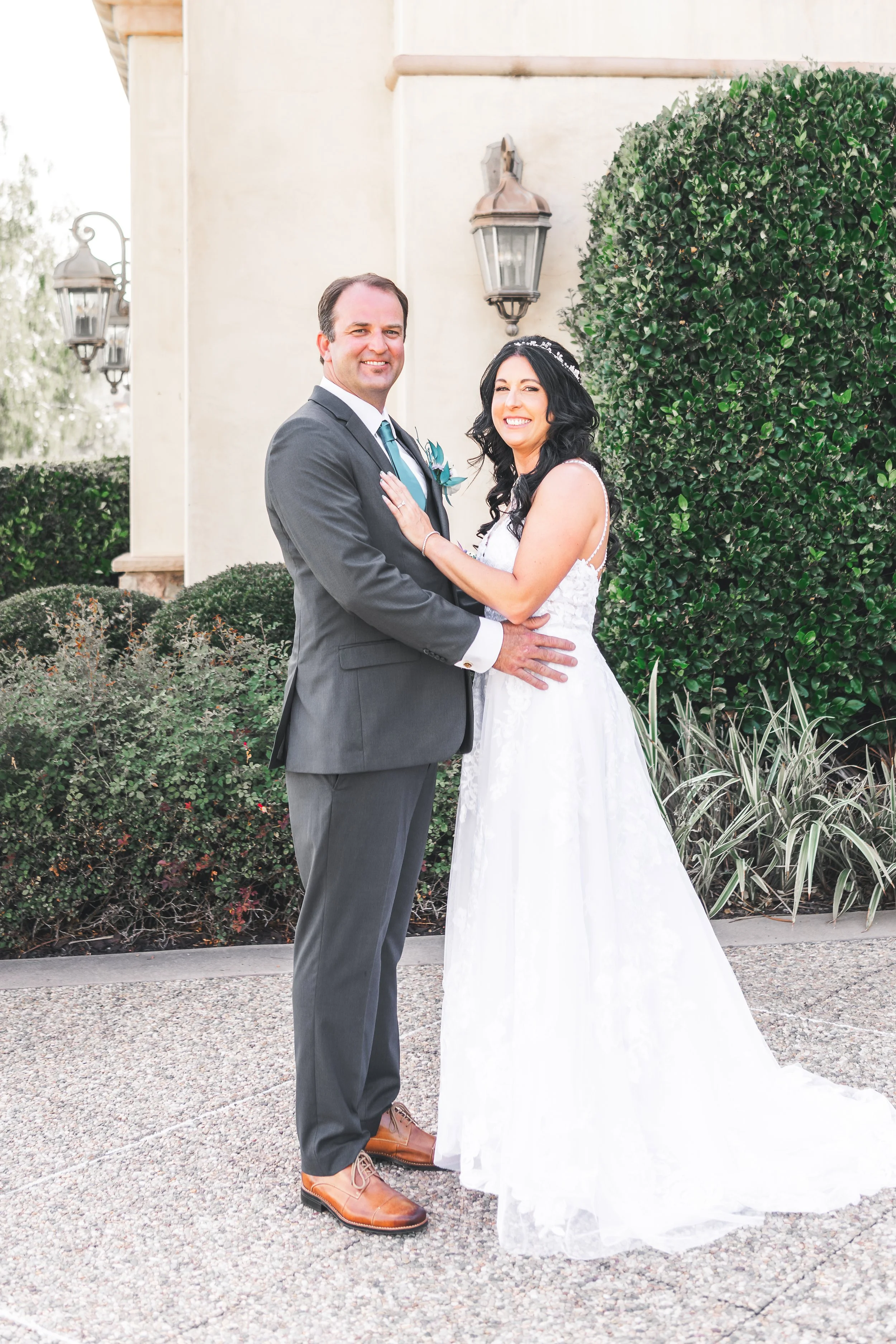 A couple posing in wedding attire outside; the groom is in a gray suit and the bride is in a white gown, standing in front of a building with decorative lamps and greenery.