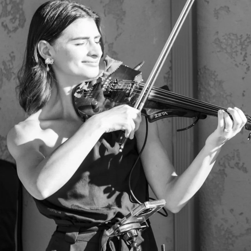Black and white photo of a woman playing an electric violin with a smile, wearing a strapless dress, outdoors.