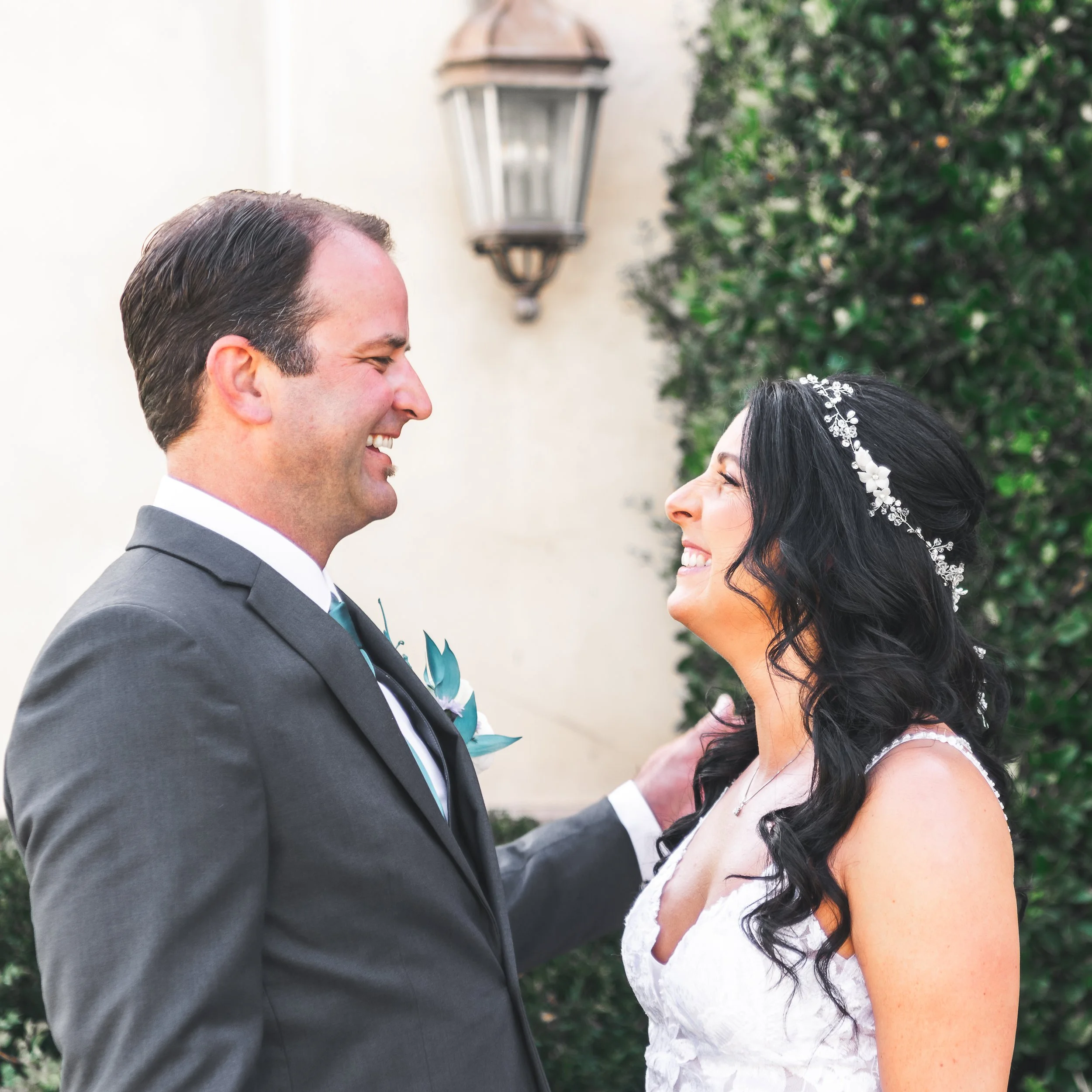 Bride and groom smiling at each other outdoors with greenery and a lantern in the background.