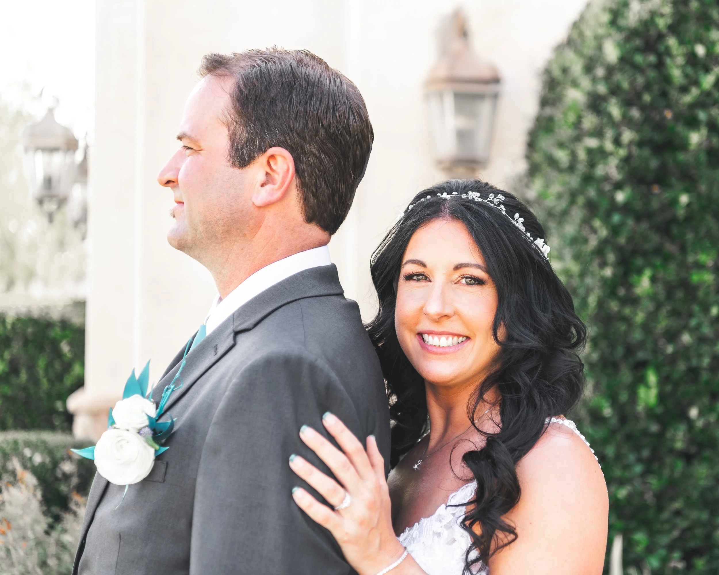 Bride and groom smiling in wedding attire, bride with long dark hair and floral headband, groom wearing gray suit with white rose boutonniere, outdoors with greenery and lanterns in background.