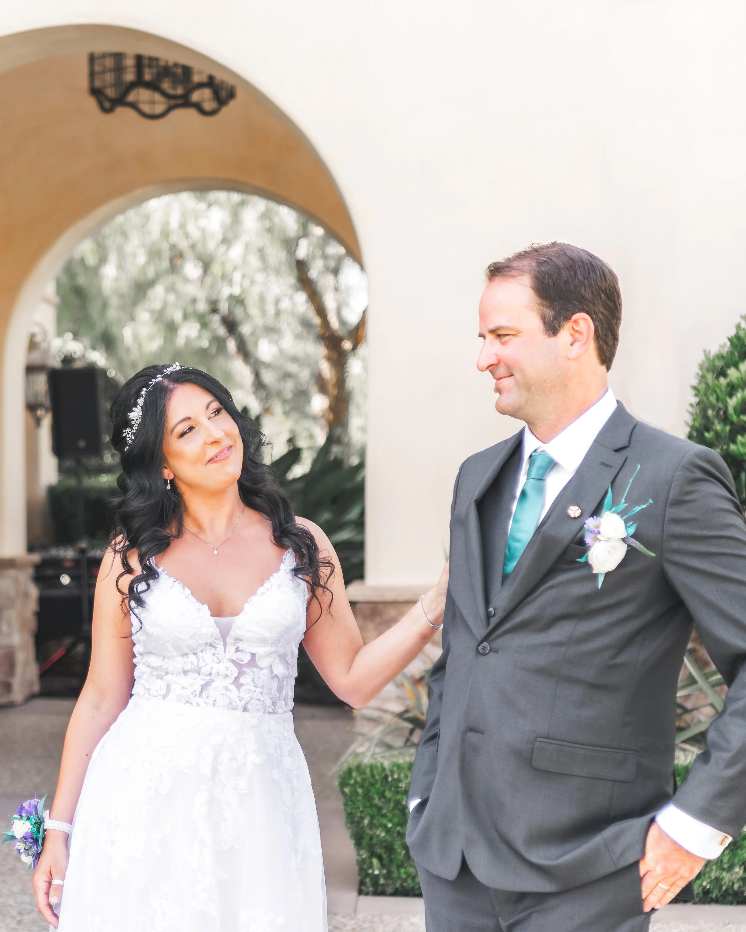 Bride and groom in wedding attire, bride wears a white dress with lace embellishments and a floral headband, groom in a gray suit with teal tie, standing outdoors by an archway.