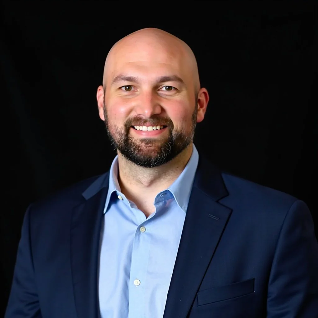 A professional headshot of a bald man with a beard, wearing a blue suit jacket and light blue dress shirt, smiling against a black background.
