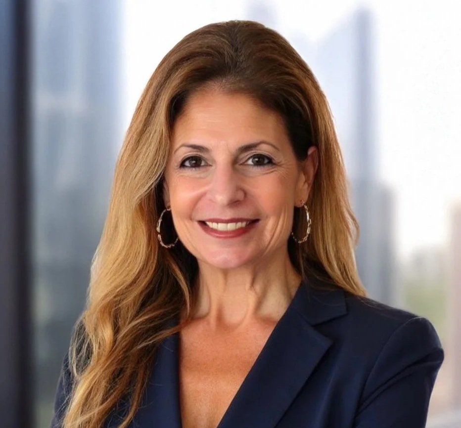 A woman with light skin, shoulder-length brown hair, and a bright smile, dressed in a dark blazer and a purple top, posing against a gray background.