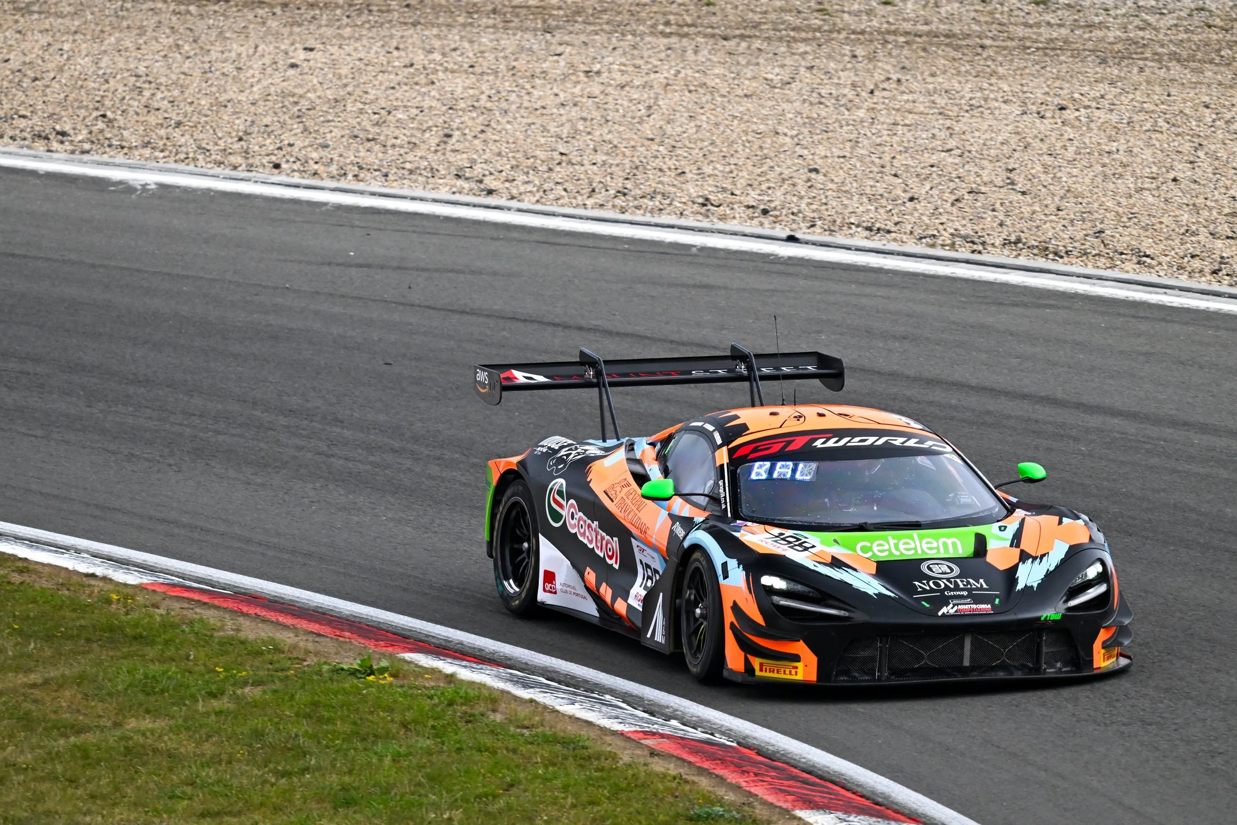 A race car with blue, orange, and black livery speeding on a racetrack with blurred surroundings.