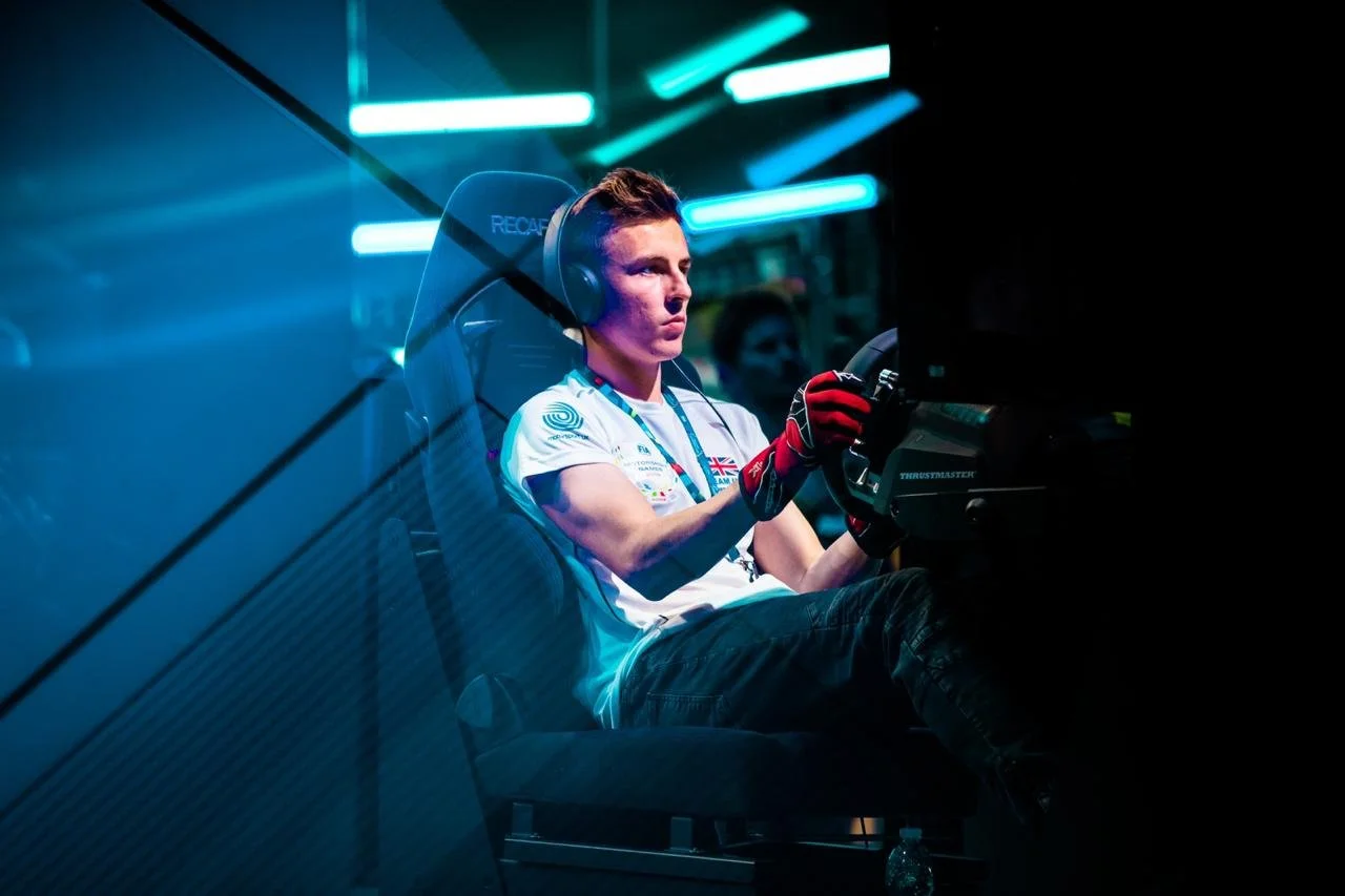 Young man in gaming chair wearing headphones and racing gloves, focused on a racing simulator game with bright blue neon lights overhead.