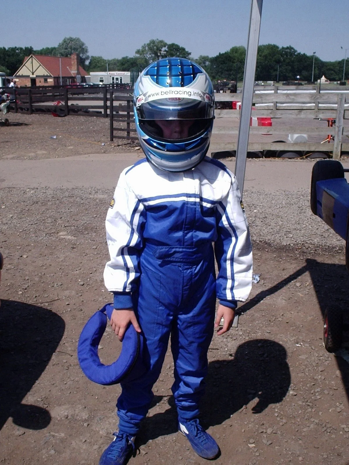 A person wearing a racing helmet and blue racing suit standing outdoors on a dirt track, holding a blue neck collar, with a race track and outdoor buildings in the background.