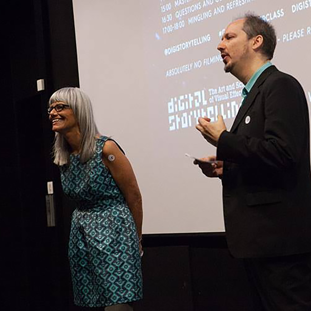 Photo of Angela Amoroso (left) and Kim Baumann Larsen (right) on stage with the program on the projection screen in the background