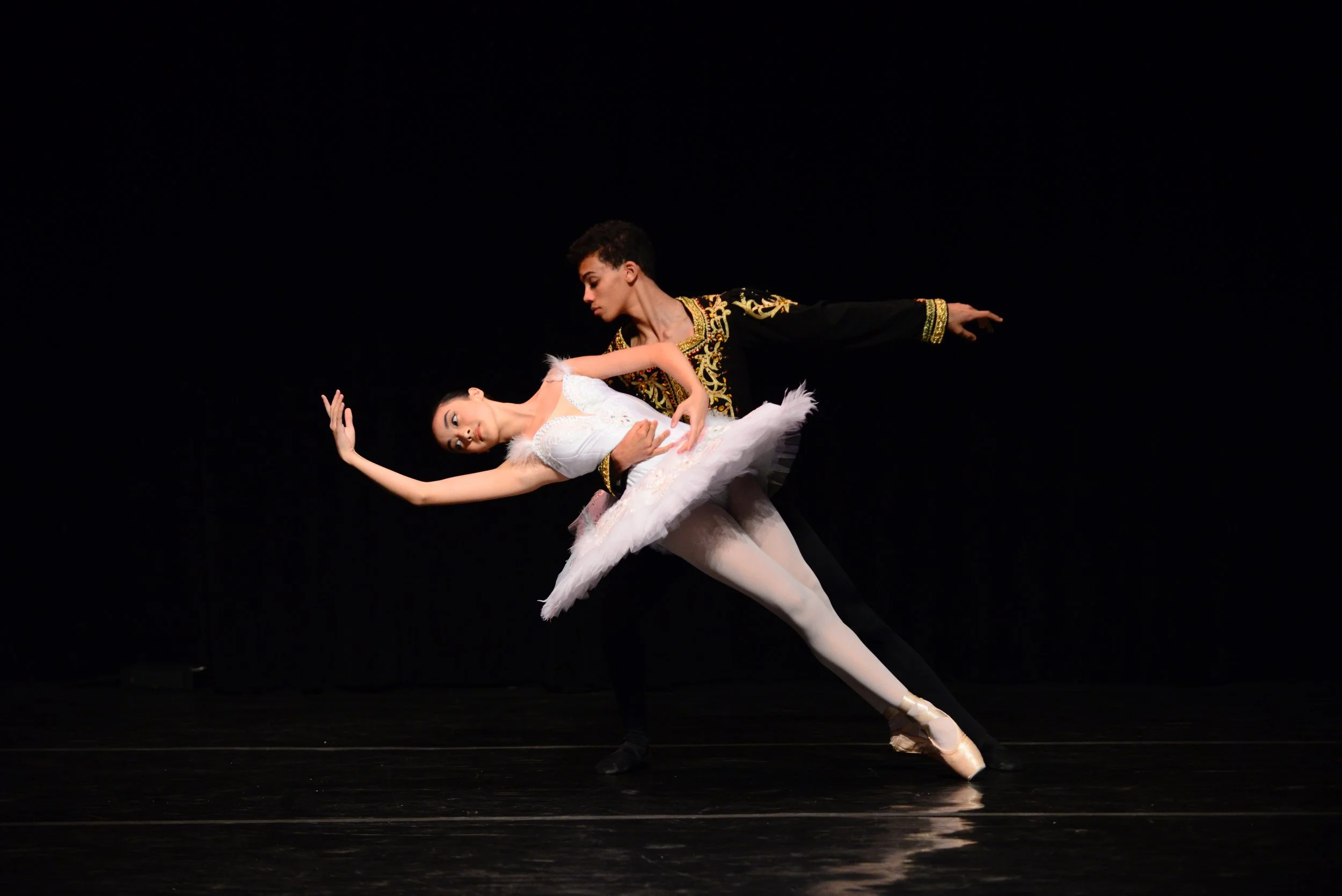 Two ballet dancers performing on stage, one male and one female, with the male supporting the female in a ballet pose, against a black background.