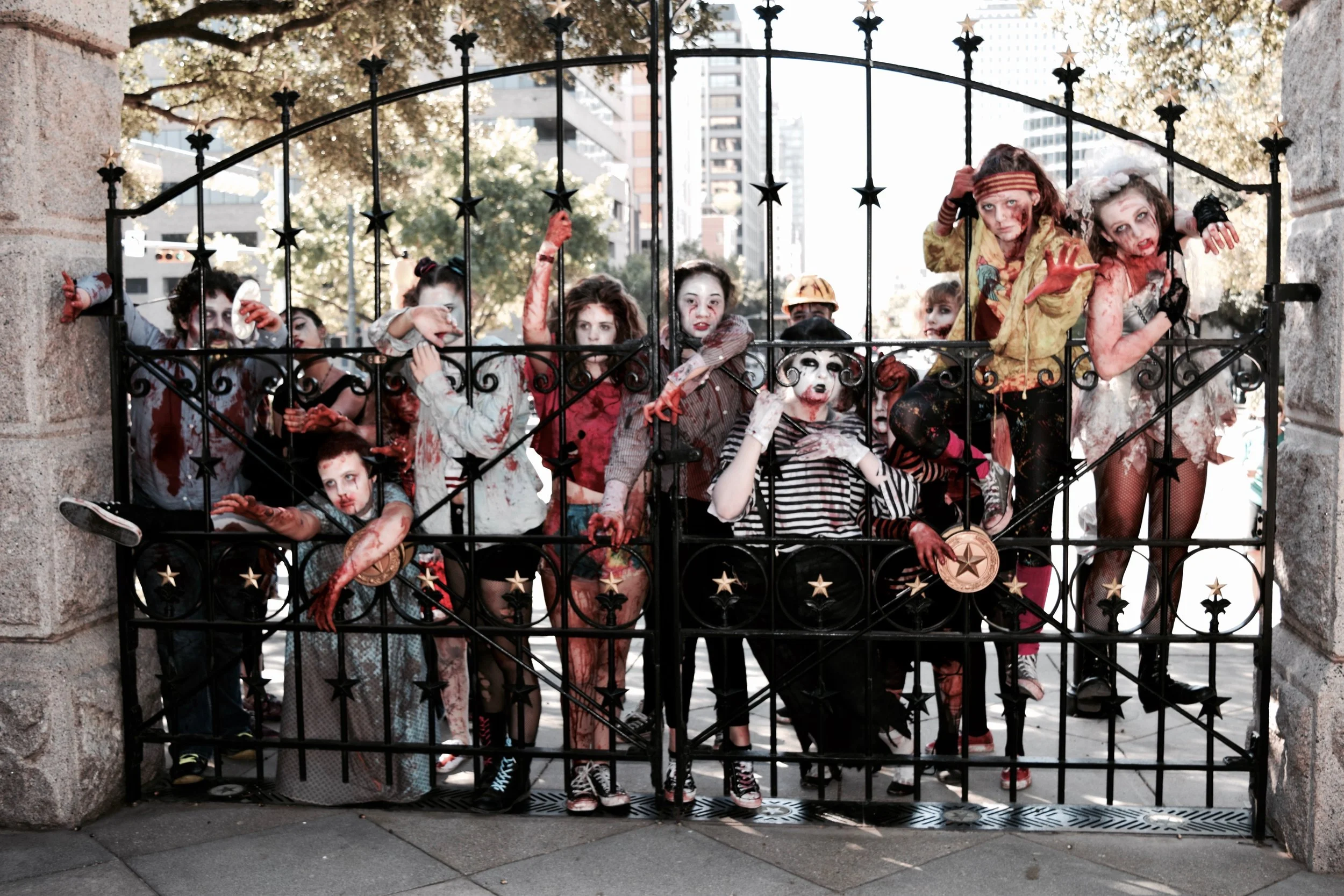 Children dressed as zombies behind a metal gate, with zombie makeup and costumes, in a city park setting.