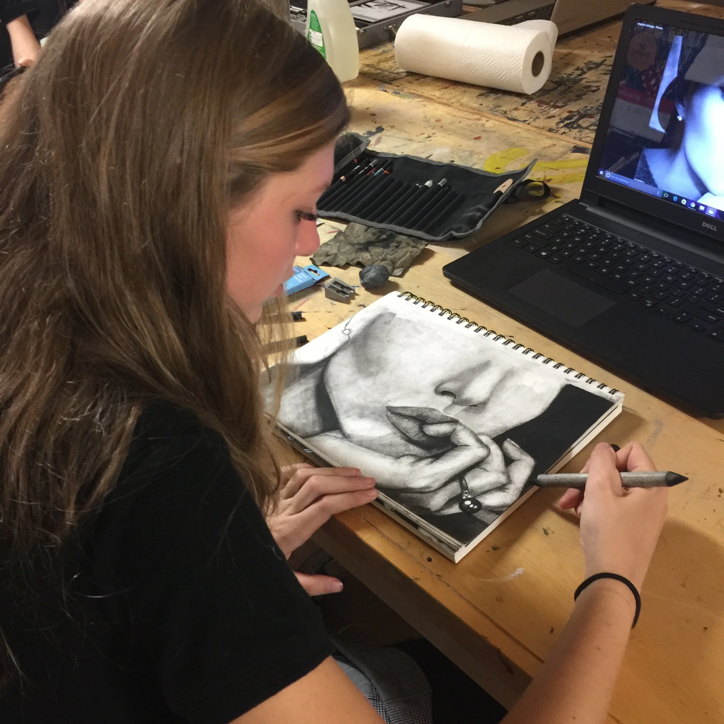 A woman drawing a realistic portrait of a woman's face and fingers on a sketchbook at a wooden table, surrounded by drawing tools, a laptop, and a paper towel roll.