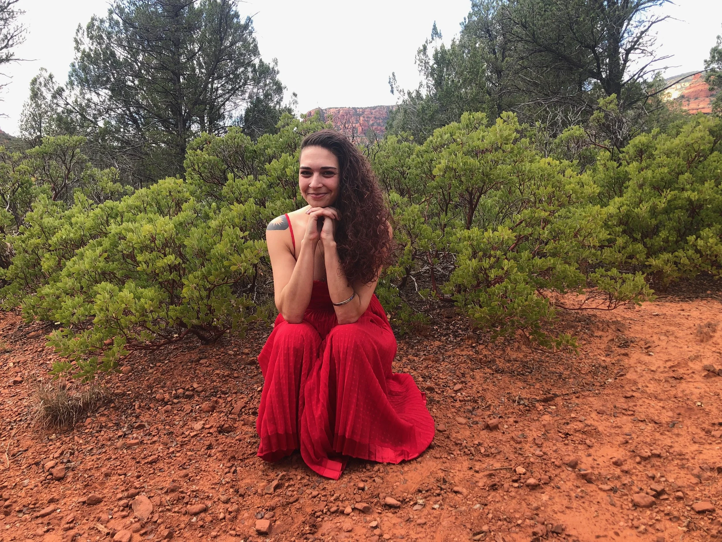 me crouched in a red dress on the red dirt of Sedona with pretty manzanitas in the background