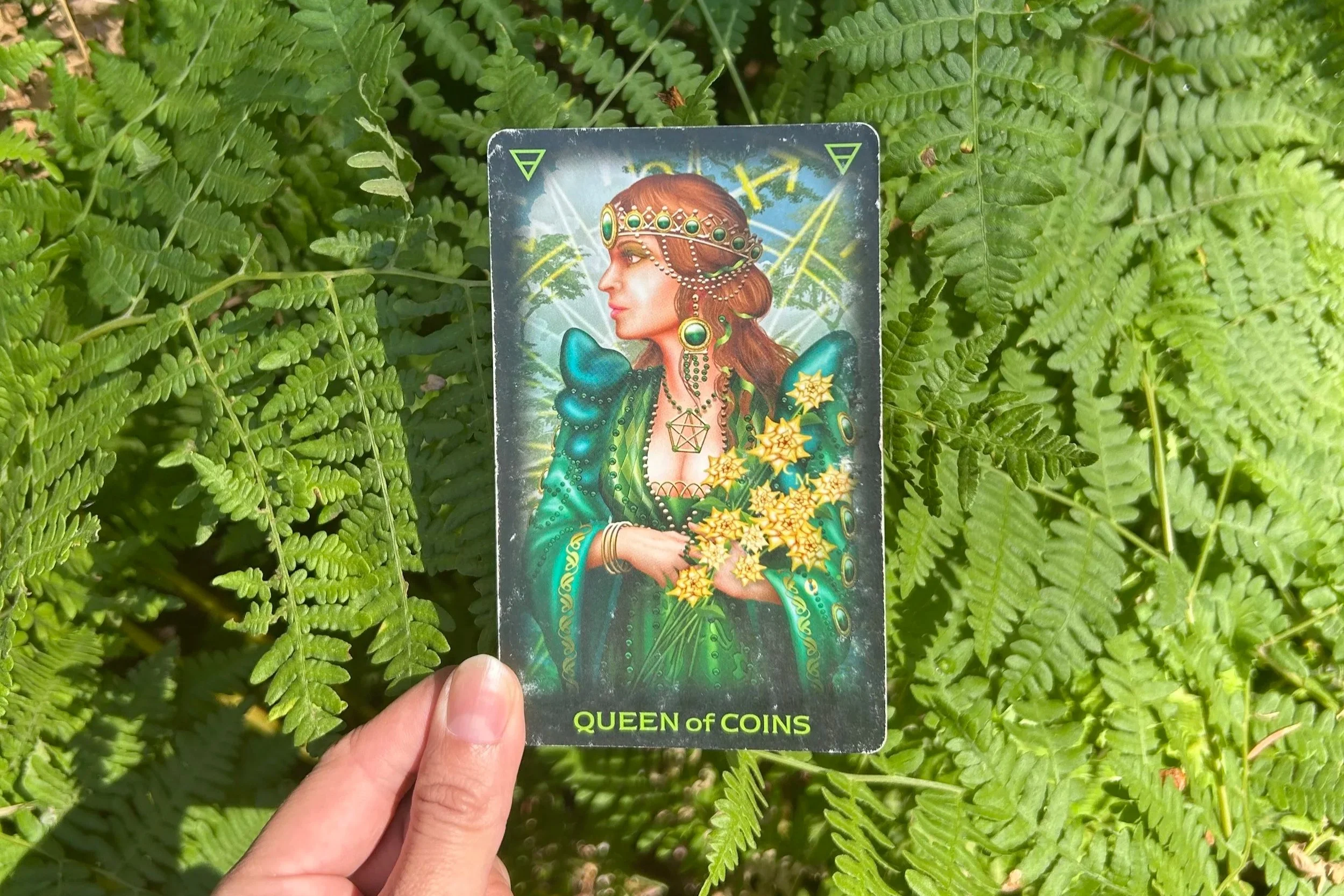 the queen of coins, the queen of pentacles, held up to a background of lush green ferns. she holds a bouquet of narcissus and looks off to her right. her necklace is a pentacle and her ears and head are adorned with jewelry made of coins.earth energy
