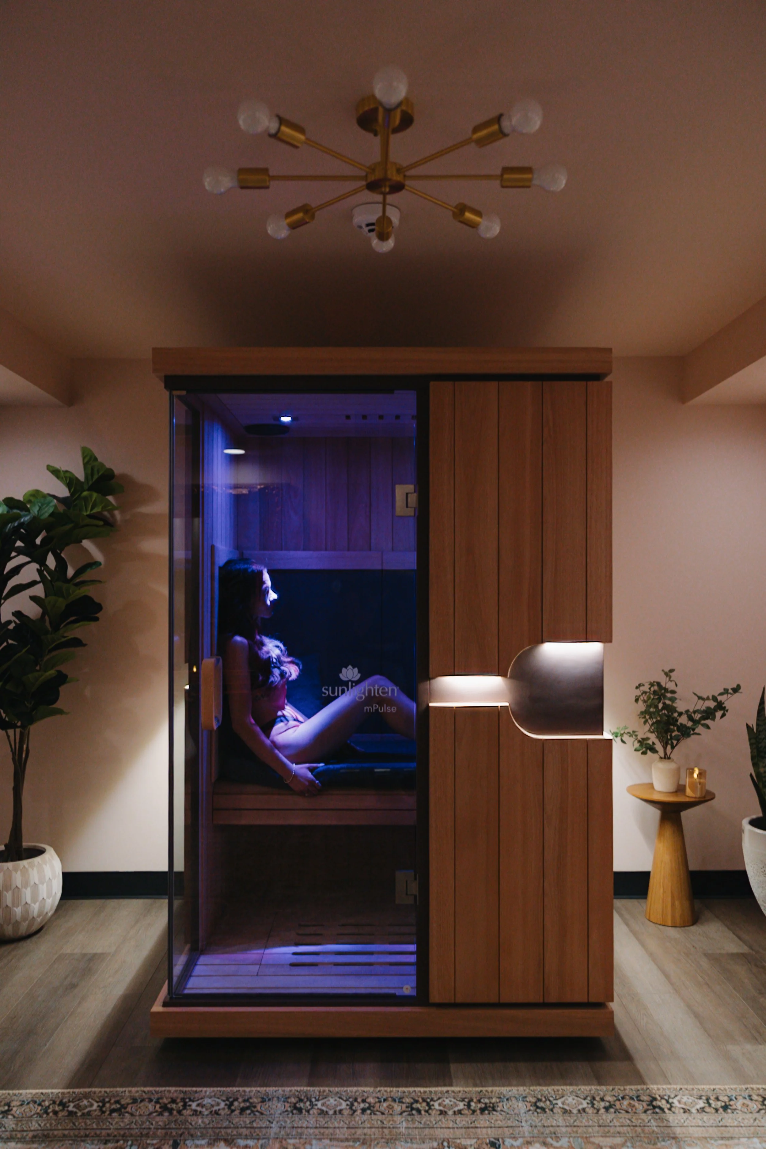A woman sitting inside a wooden and glass infrared sauna, illuminated by purple light, in a modern living space with plants, a side table, and a ceiling light fixture.