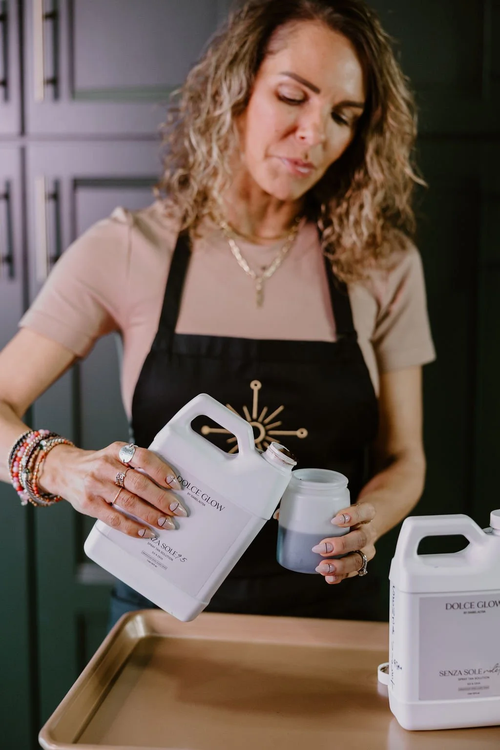 Woman pouring powder from a white container into a small jar, standing in front of a tray.