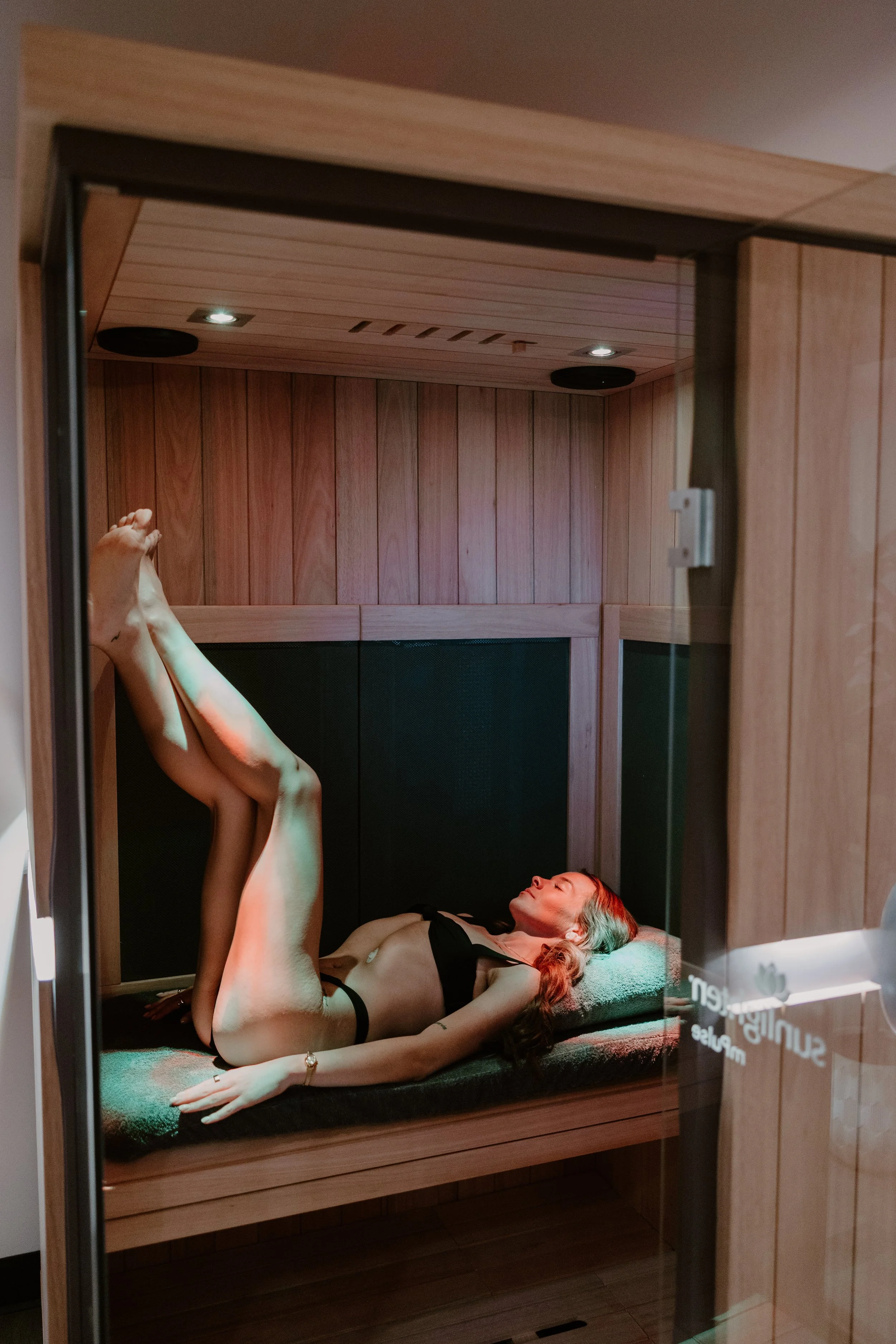 A woman lies inside a wooden sauna, wearing black lingerie, with her eyes closed and one leg raised against the wall, in a relaxed pose.