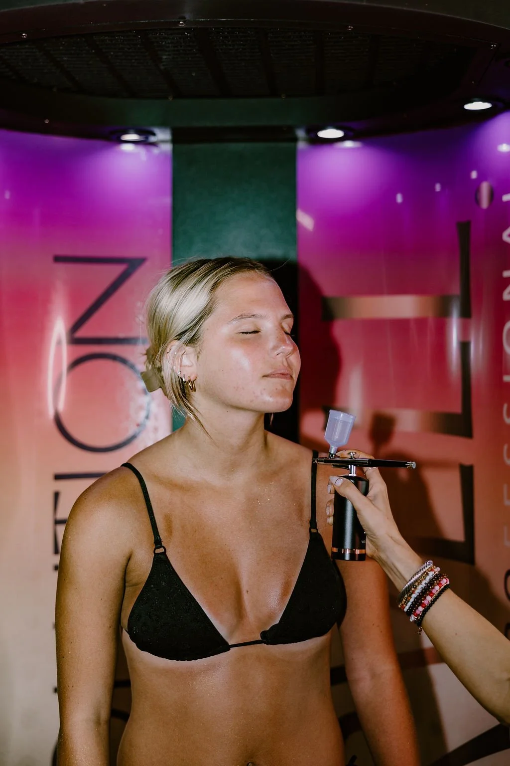 A young woman with blonde hair and earrings relaxing with her eyes closed as a UV skin test is being performed with a handheld device at a wellness or skincare facility.