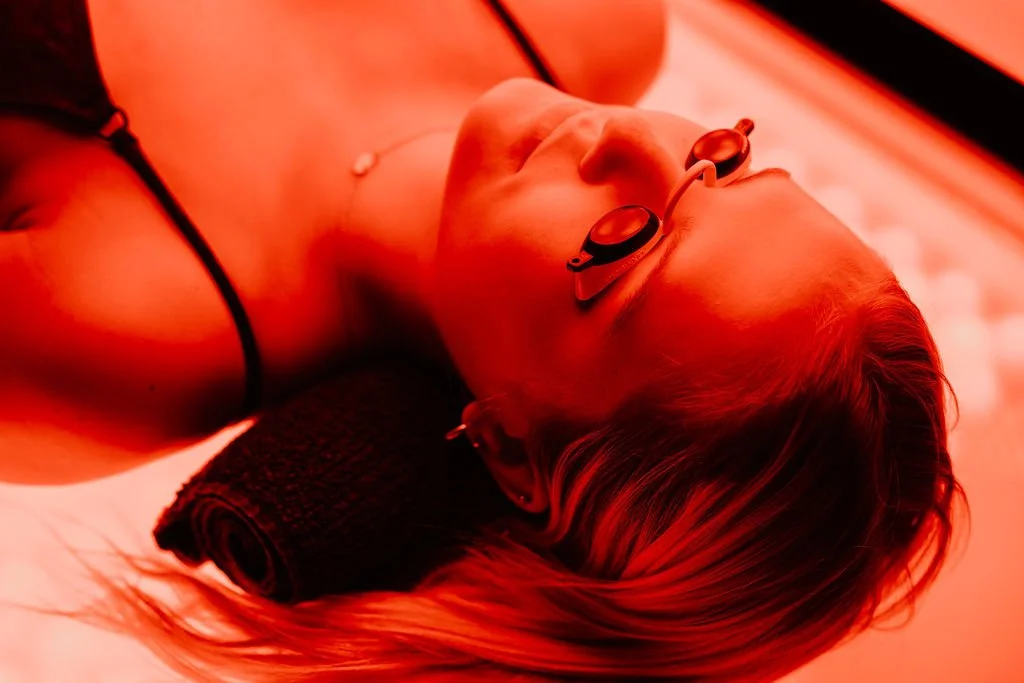 A woman lying down on a massage table, wearing goggles and a swimsuit, with a towel rolled under her neck, in a room illuminated with red lighting.