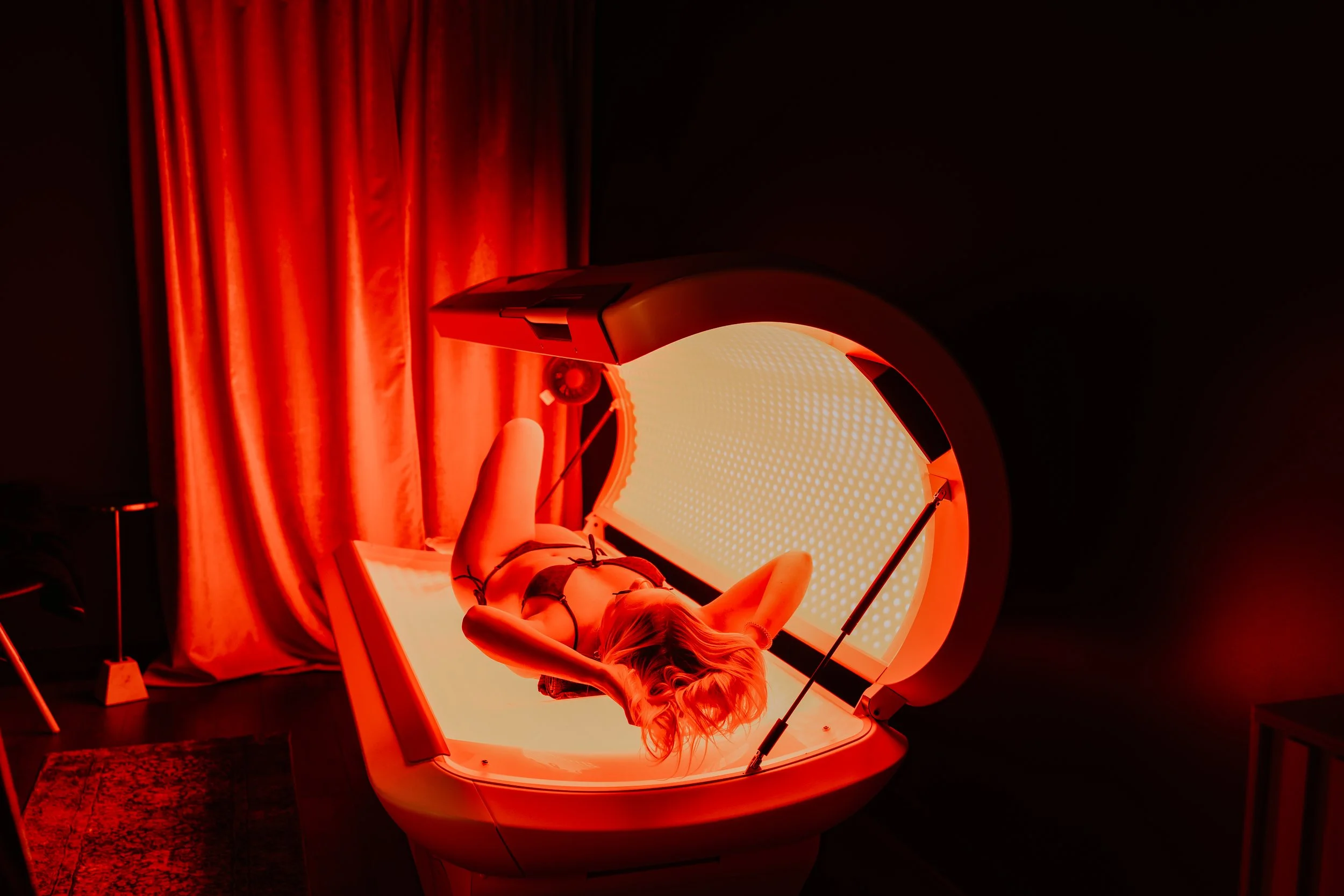 A woman lying on her back inside a red light therapy bed with a large circular light emitting red illumination, surrounded by dark room and red curtains.