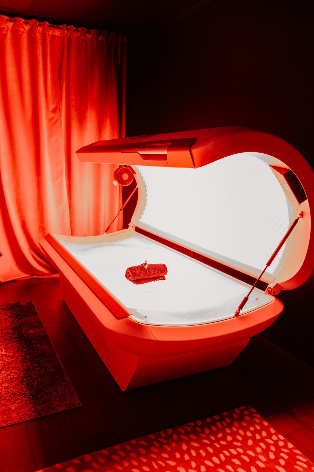 A red tanning bed with a towel on it, illuminated under red lights, in a room with red curtains and patterned rugs.