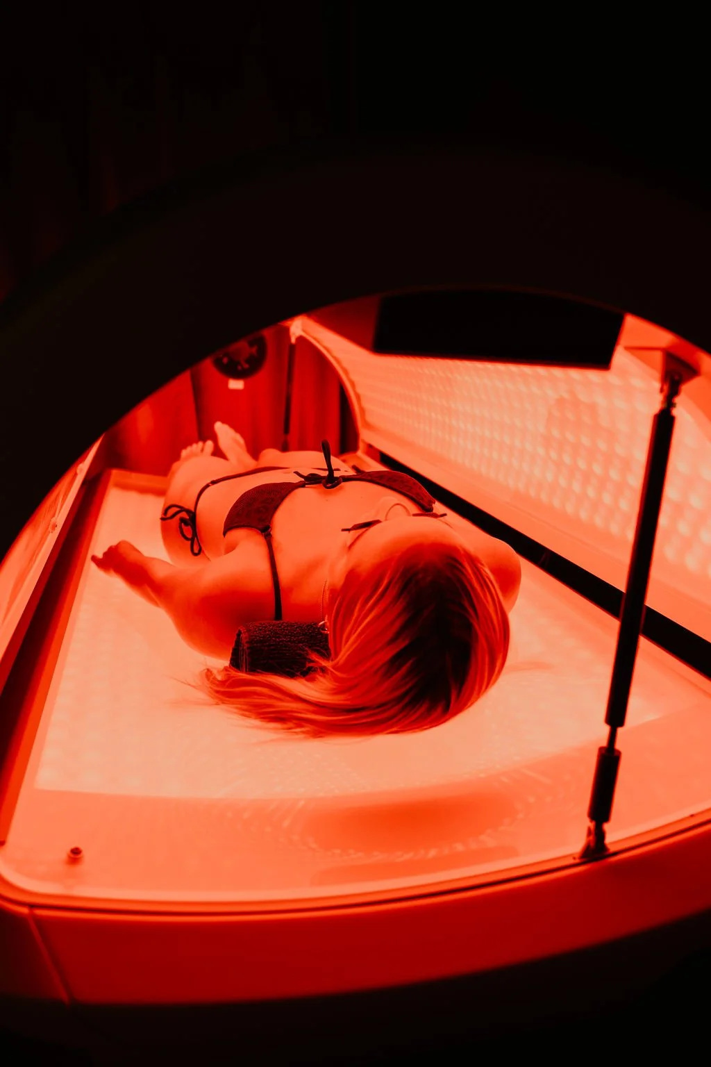 A woman lying on a bed inside a red light therapy or tanning bed, wearing black swimwear.