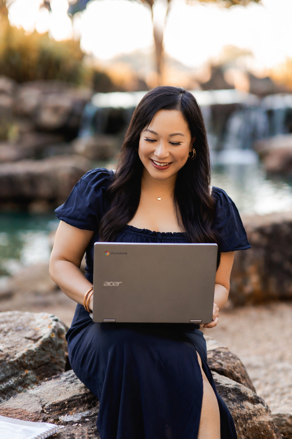 A woman in a navy dress sitting on rocks by a river with a small waterfall, smiling while looking at her laptop.