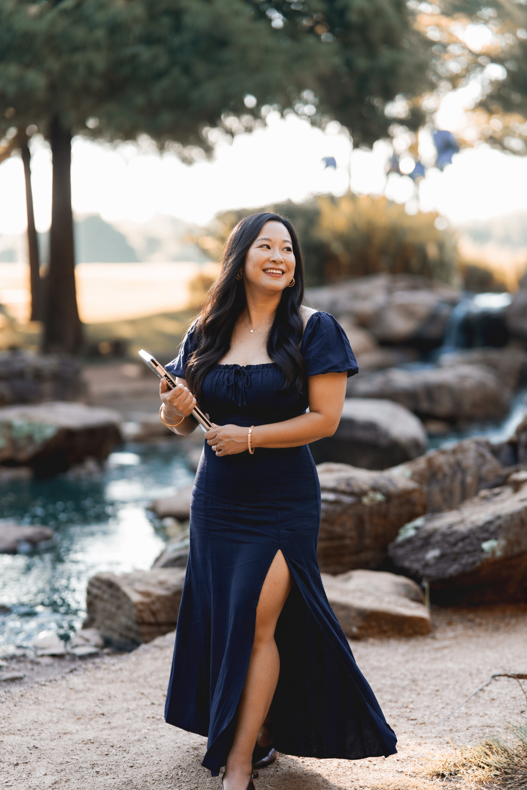 A woman wearing a navy blue dress with a high slit and puffed sleeves standing outdoors near a creek with rocks and trees, smiling and holding a smartphone.