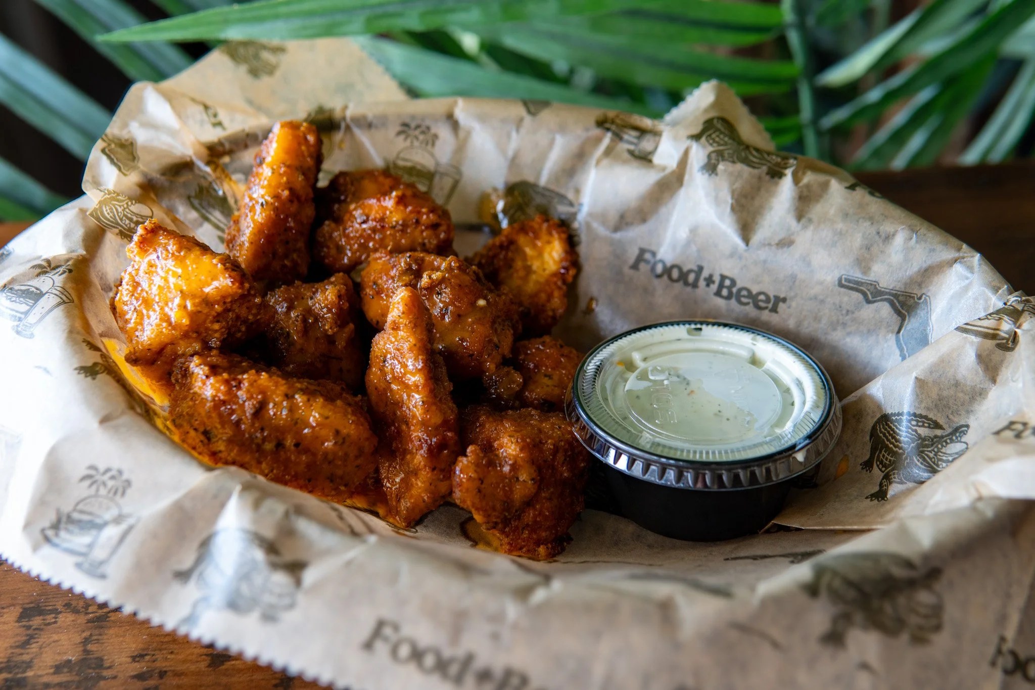 A basket of fried chicken wings coated in sauce, served with a side of creamy dipping sauce in a small container, on food wrap with printed design.