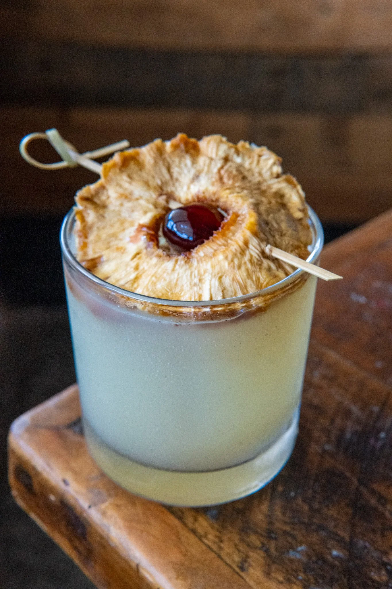 A creamy beverage topped with a dried pineapple ring and a maraschino cherry, served in a glass on a wooden tray.