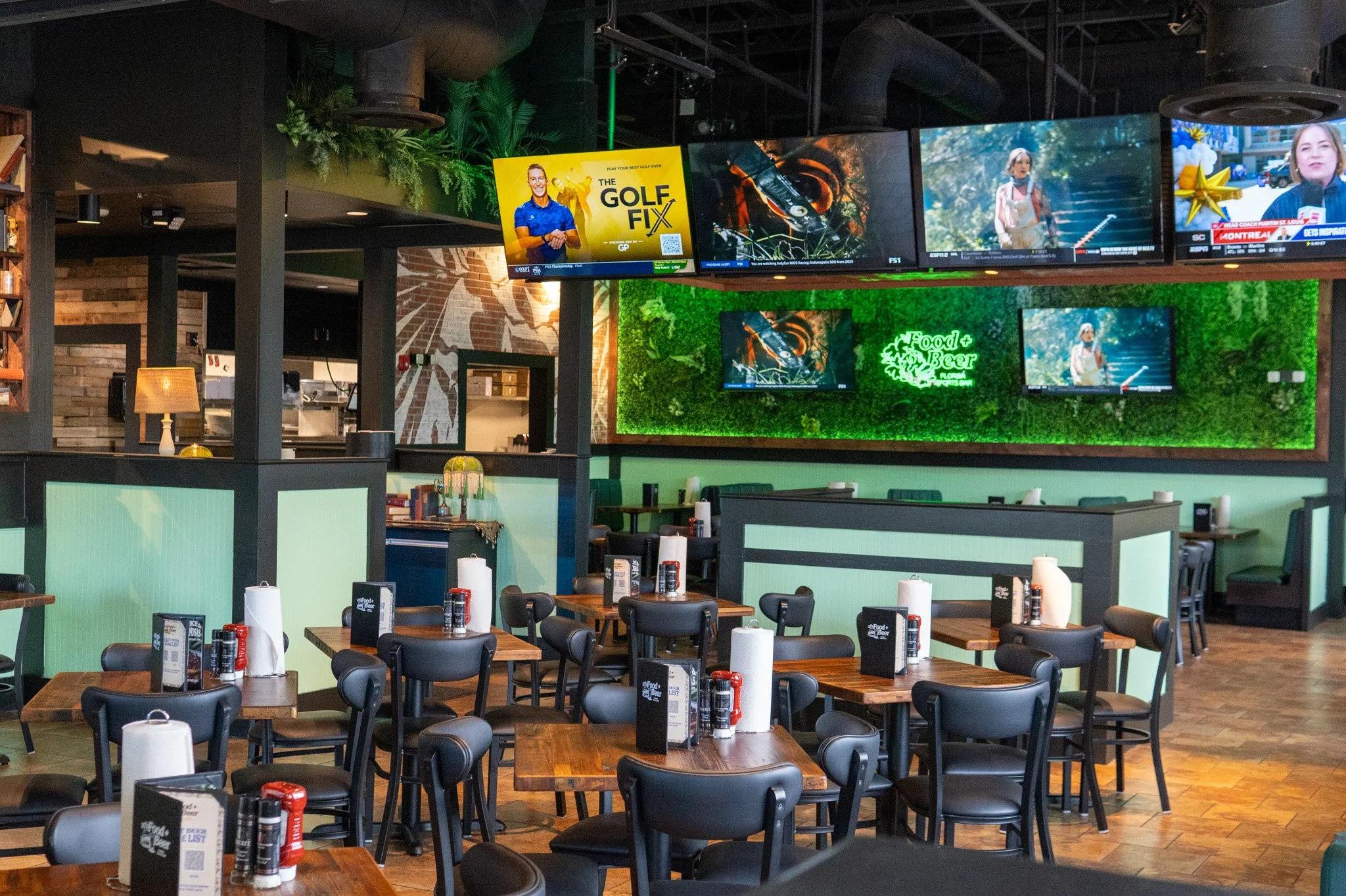 Interior of a restaurant with wooden tables, black chairs, multiple television screens showing sports and advertisements, a green wall with a neon sign that says 'Food + Beer', and some plants and decorative lights.