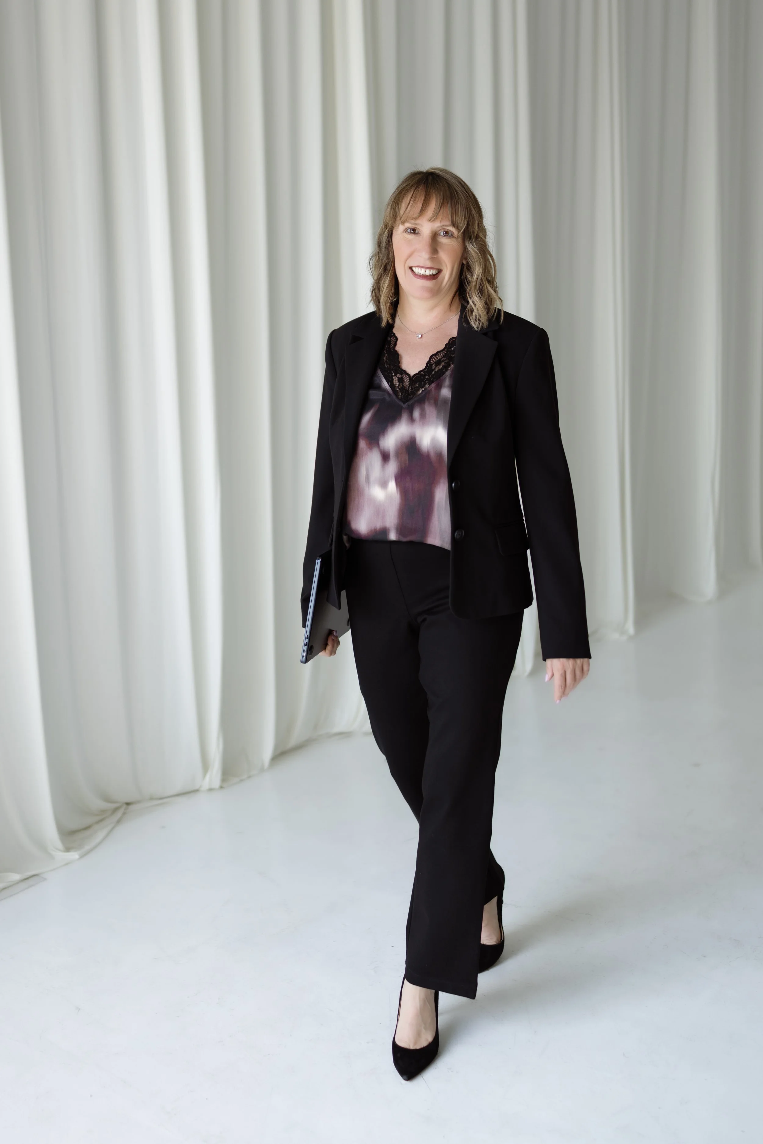 Jen Ostrich walking on a white floor in front of white curtains, wearing a black blazer, black pants, and black high heels, holding a tablet in her right hand and smiling.