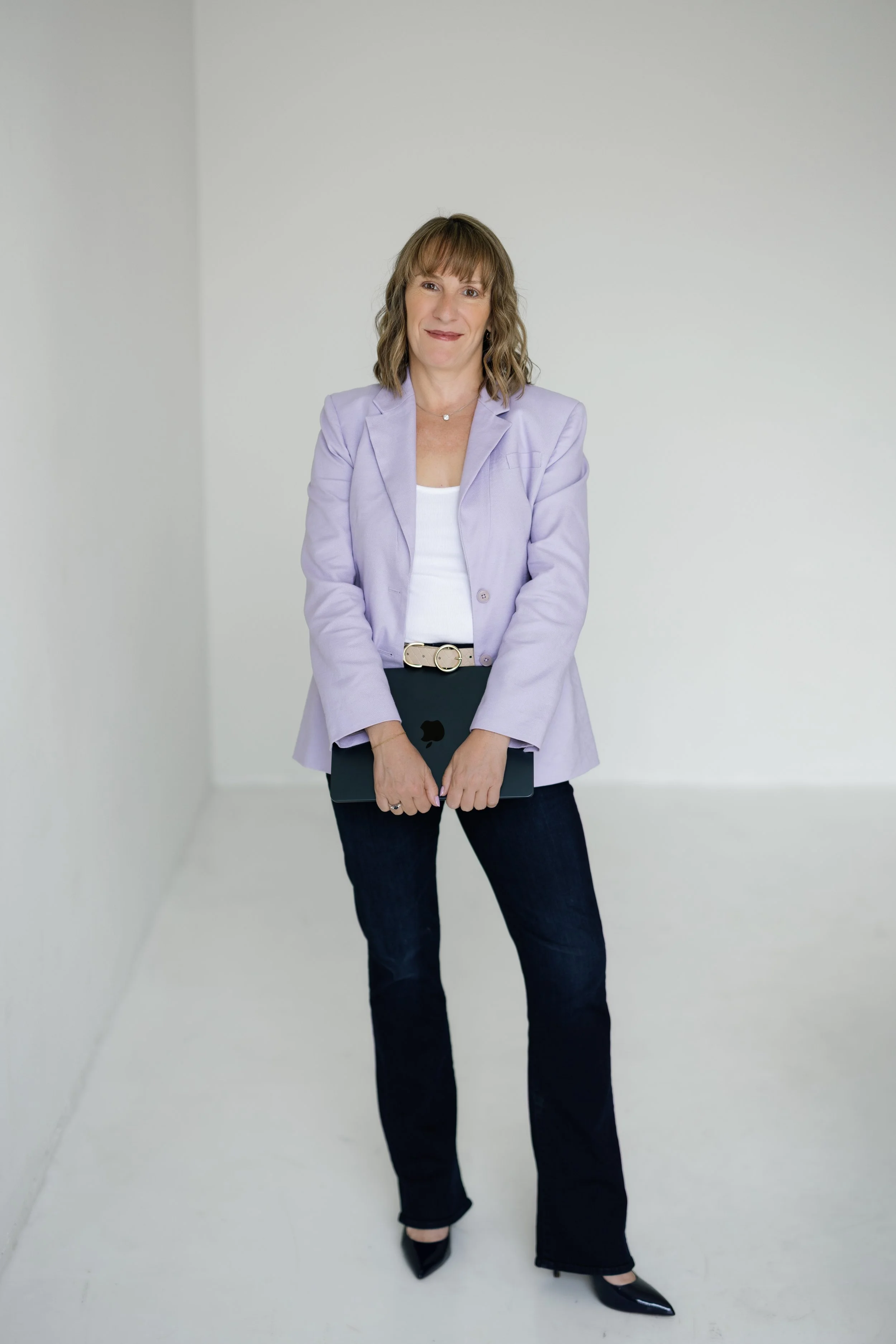 Jen Ostrich standing in a white room, wearing a light purple blazer, white top, dark pants, and black pointed-toe heels, holding a closed laptop.