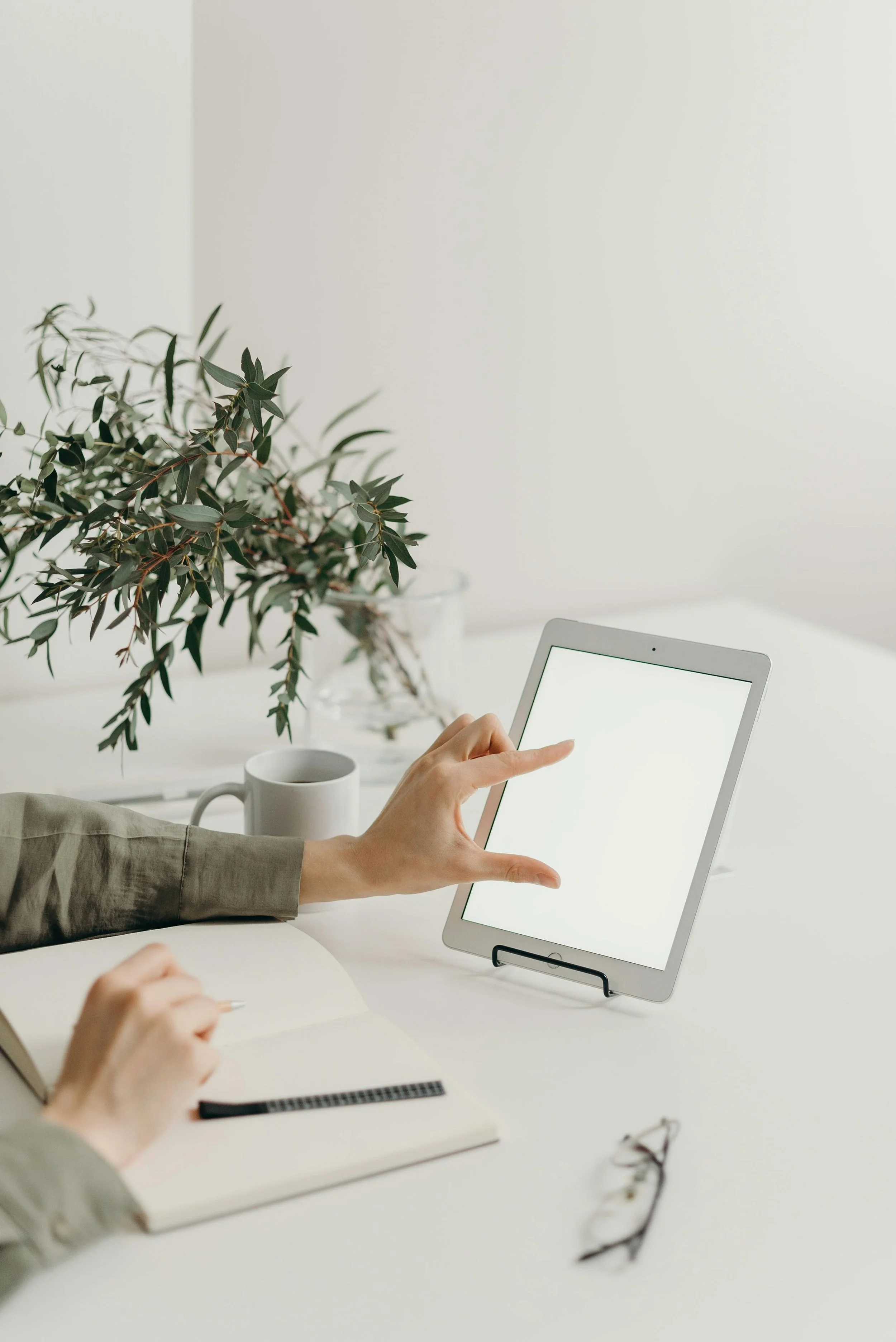 Person using a tablet on a white desk with a notebook, a cup, and glasses nearby, and a plant in the background.