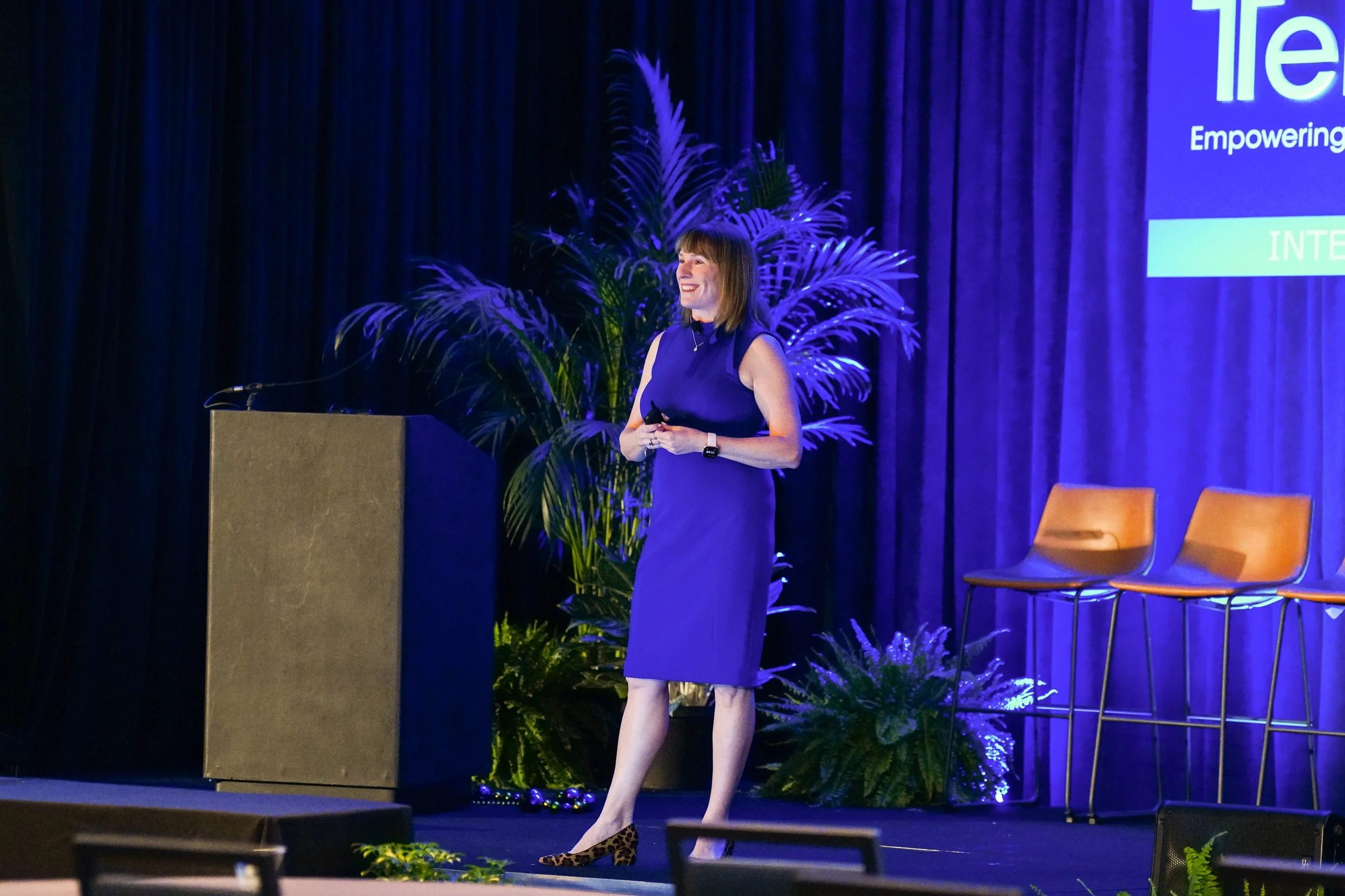 Jen Ostrich in a blue sleeveless dress standing on stage at a conference, smiling, holding a remote control, and giving a keynote at a national sales training conference on shifting your perspective.