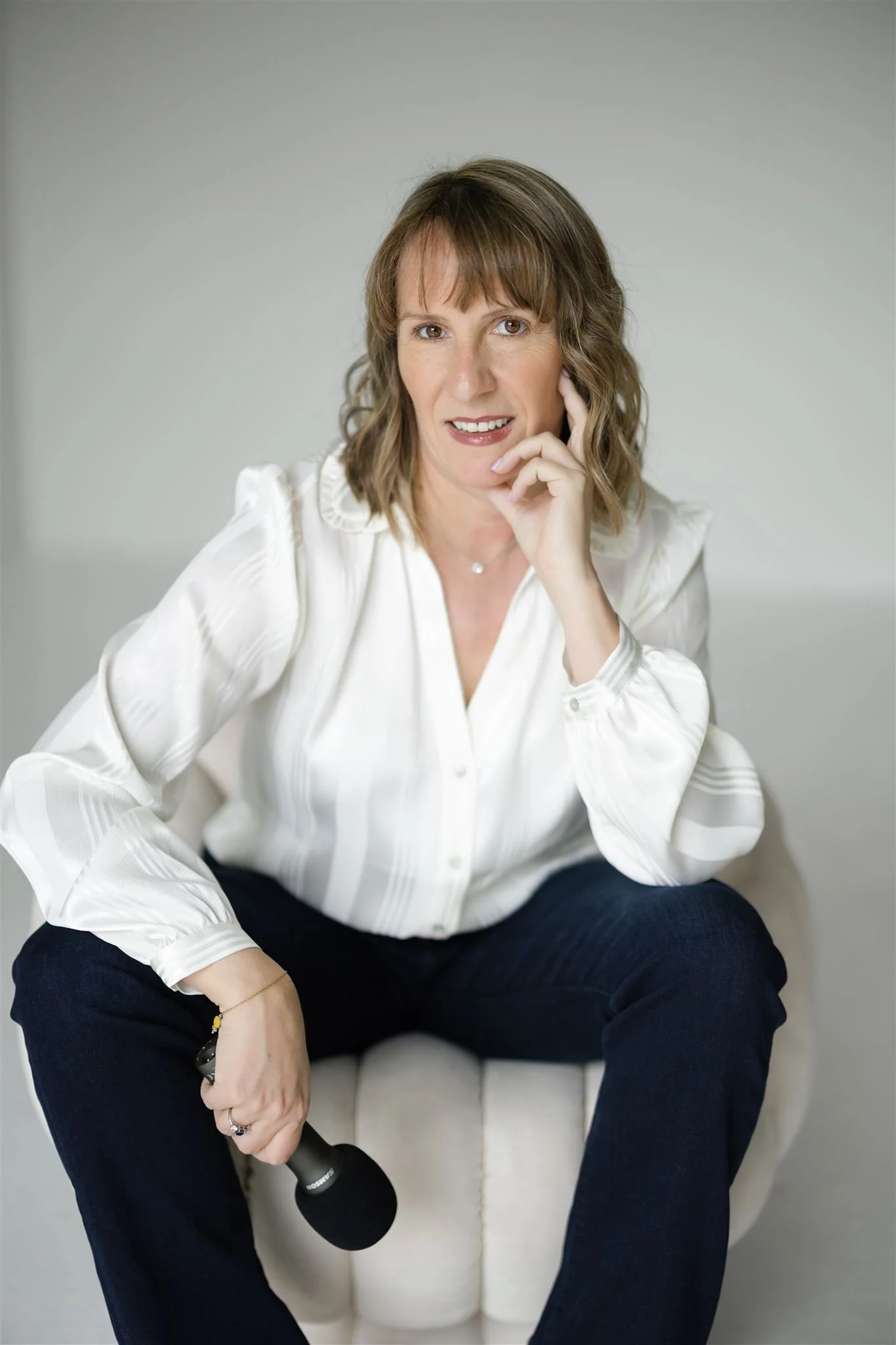 Jen Ostrich with shoulder-length light brown hair, wearing a white blouse and dark pants, sitting on a white tufted chair, holding a black microphone, with her right hand resting against her face.