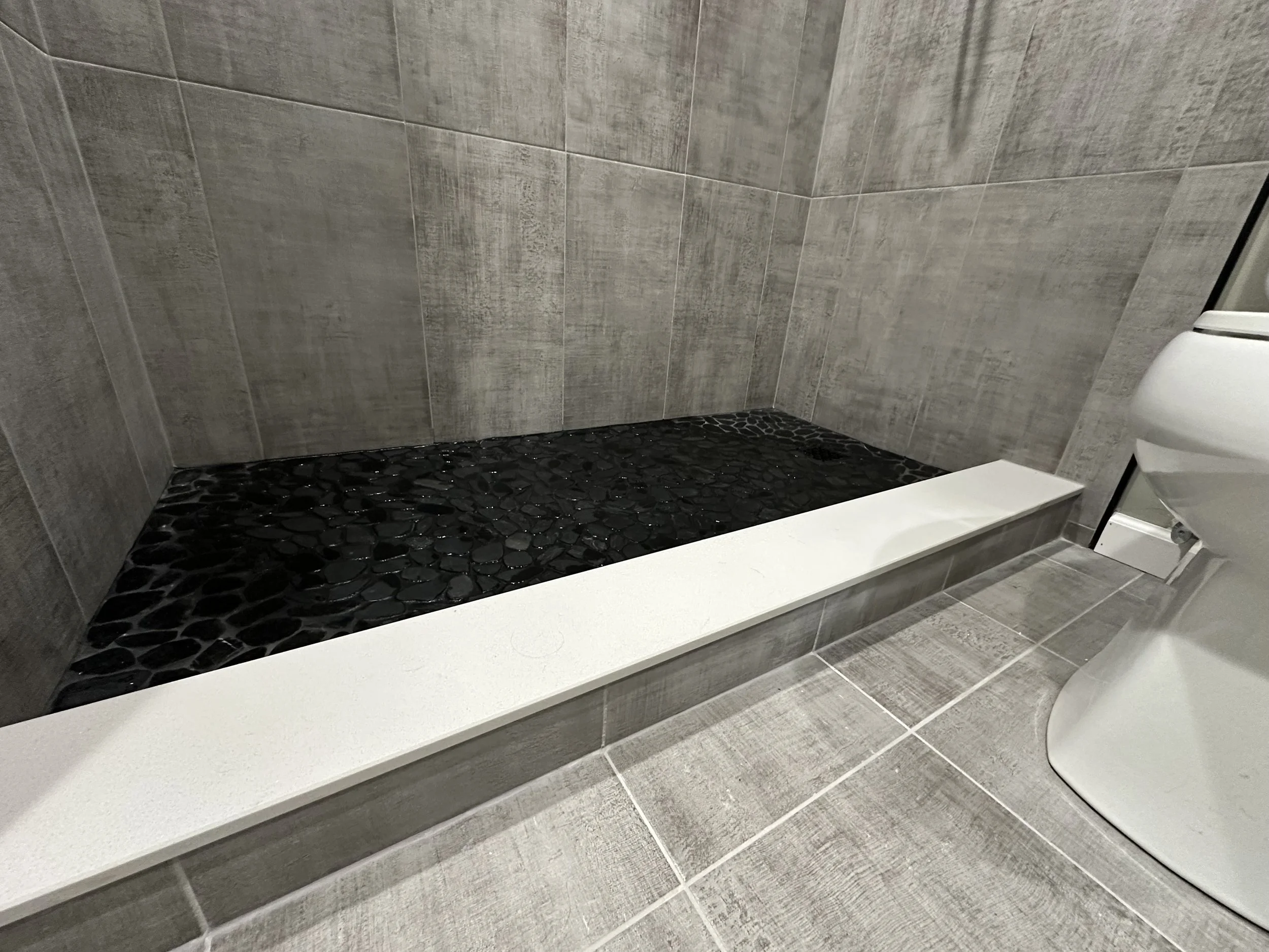 Close-up of a modern bathroom shower area with black pebble tiles, gray tiled walls, and a gray tiled floor, next to a white toilet.