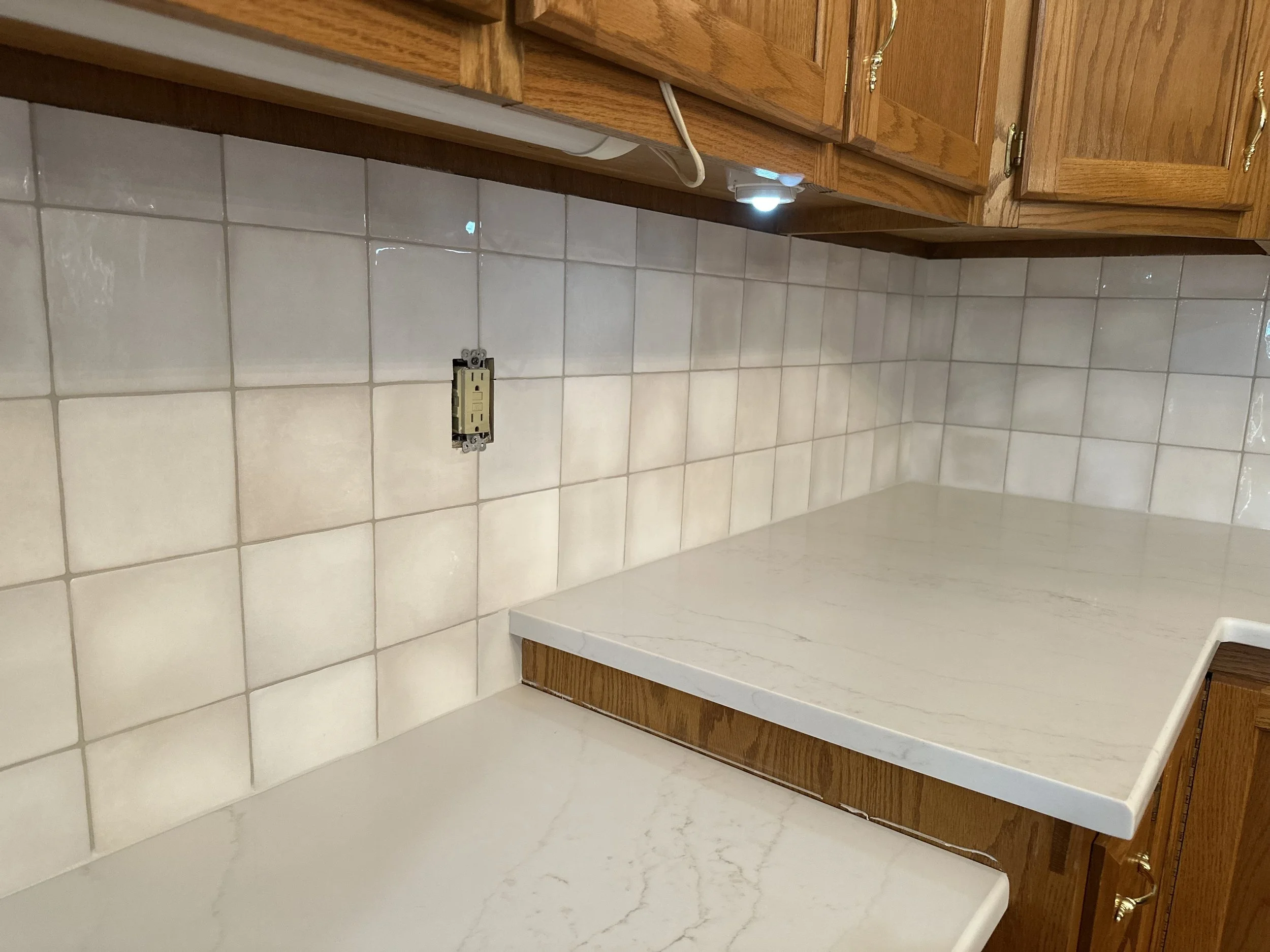 Kitchen countertop with beige tile backsplash, wooden cabinets above, electrical outlet missing cover, and under-cabinet light.