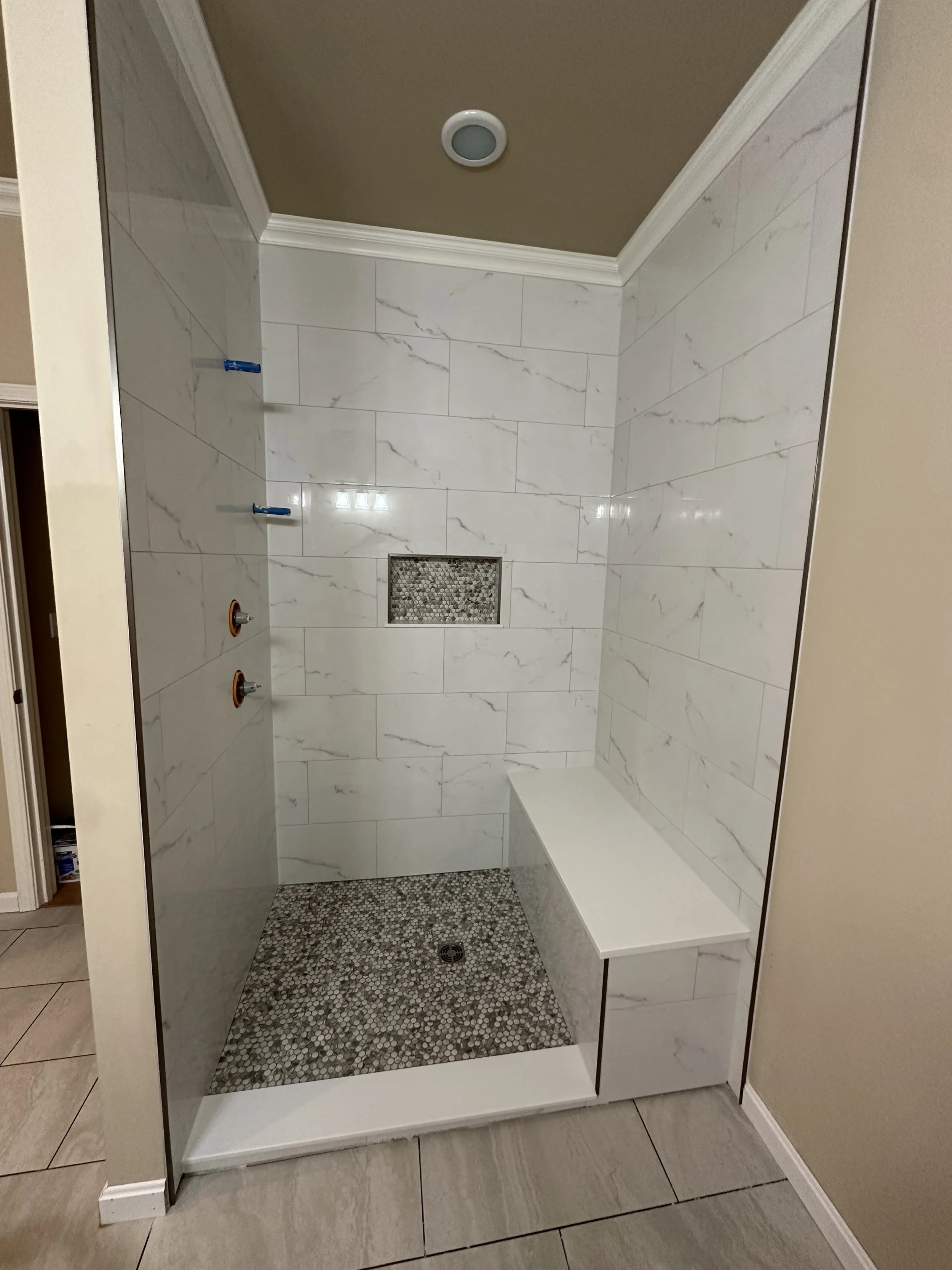A modern walk-in shower with white marble tiles, a built-in marble bench, a small niche with mosaic tiles, and pebble stone flooring.