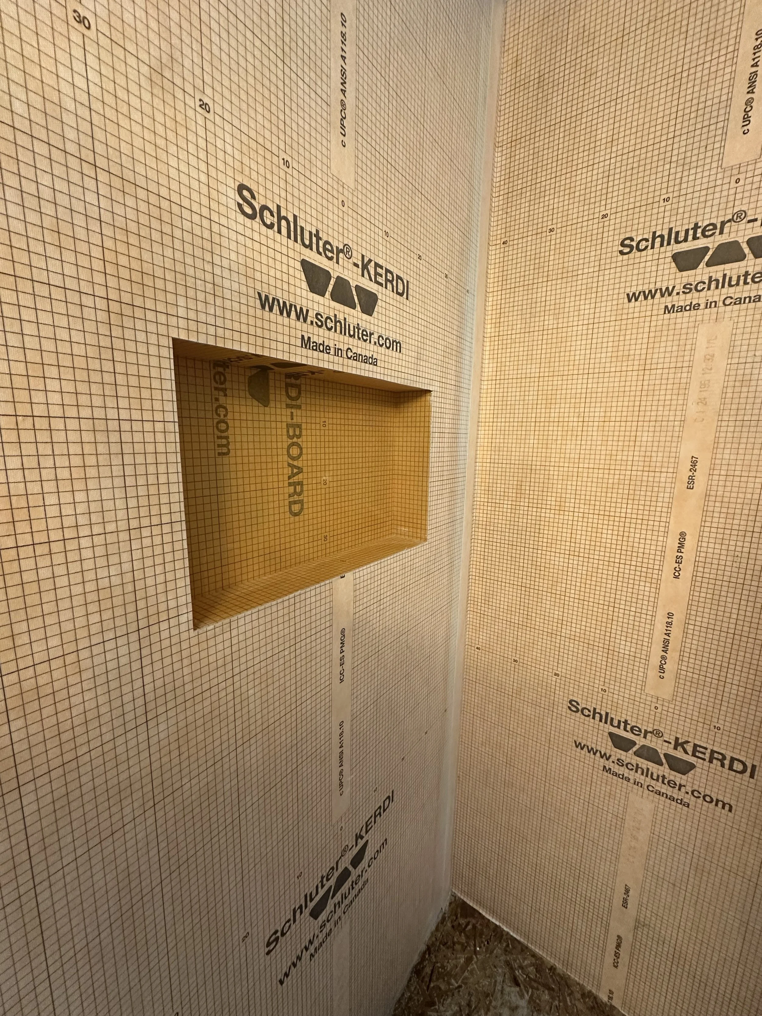 Close-up of a wall with green sheathing panels branded Schluter-KERDI, showing a rectangular cutout area and grid lines for waterproofing membrane installation.