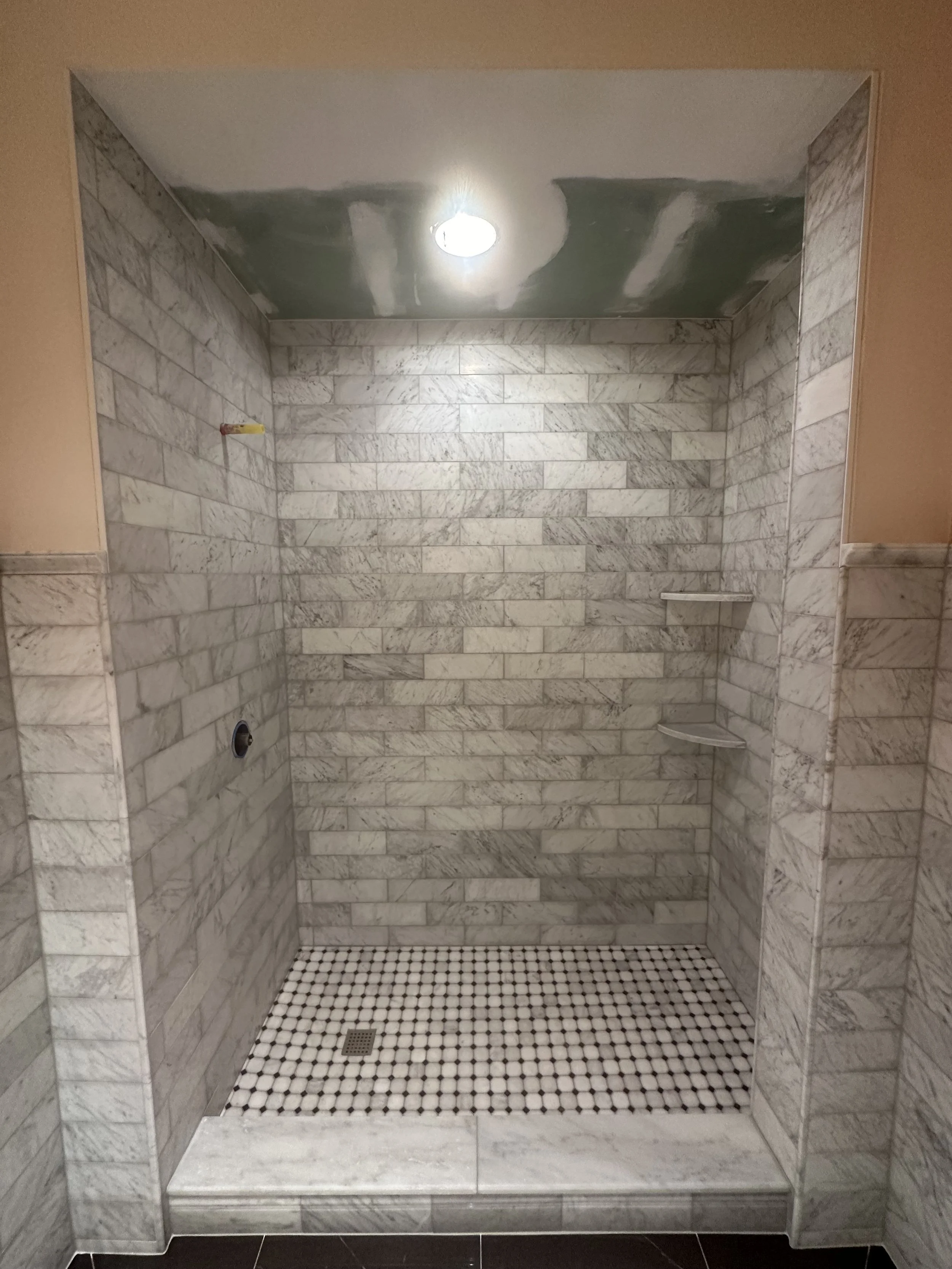 Empty marble-tiled walk-in shower with built-in corner shelves, a circular drain, and an overhead light, with beige and black tile on the floor and walls.
