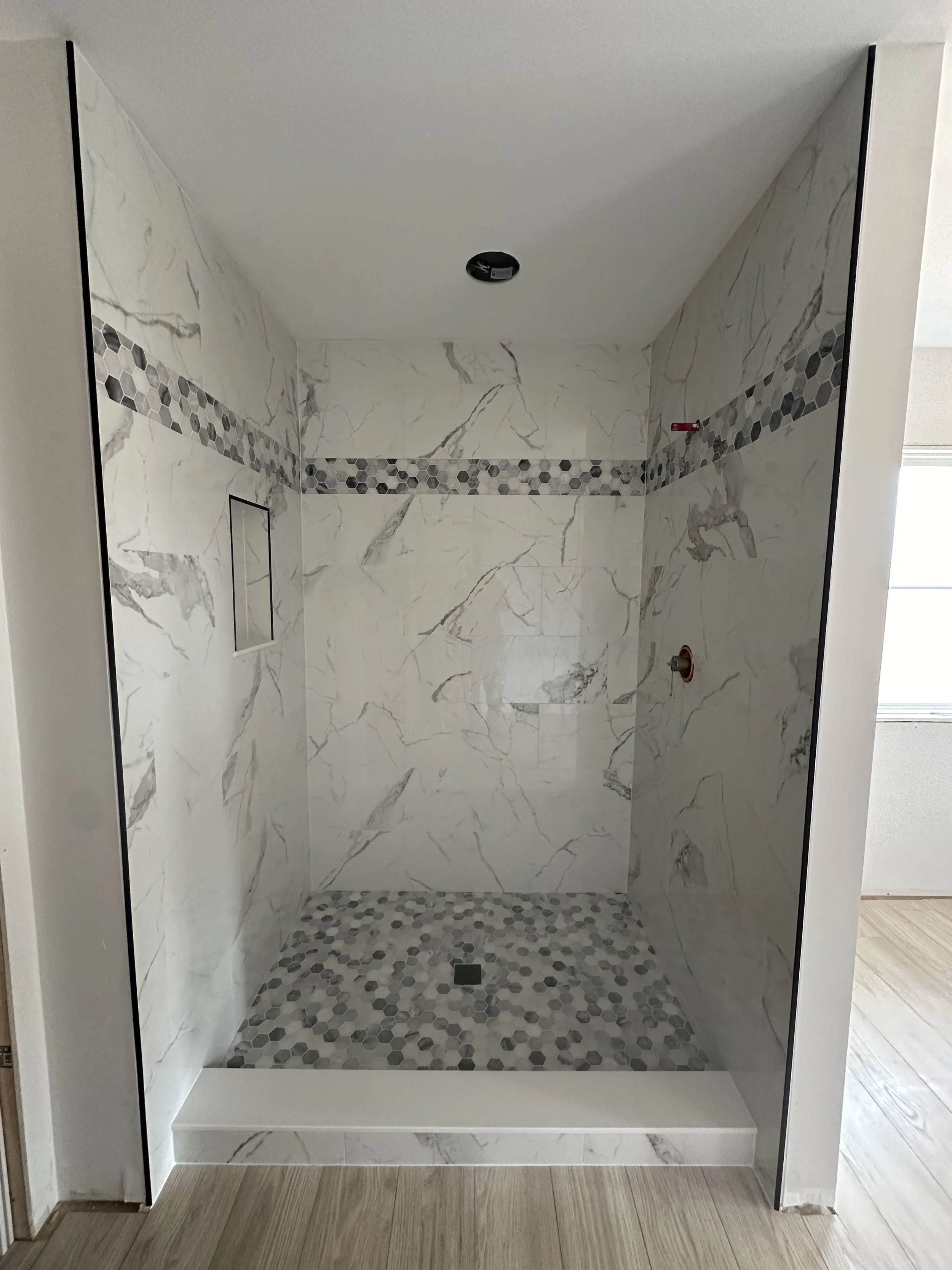 Unfinished walk-in shower with white marble walls, gray and white hexagonal pebble floor, a small niche, and a row of gray hexagonal tiles across the walls. No fixtures installed.