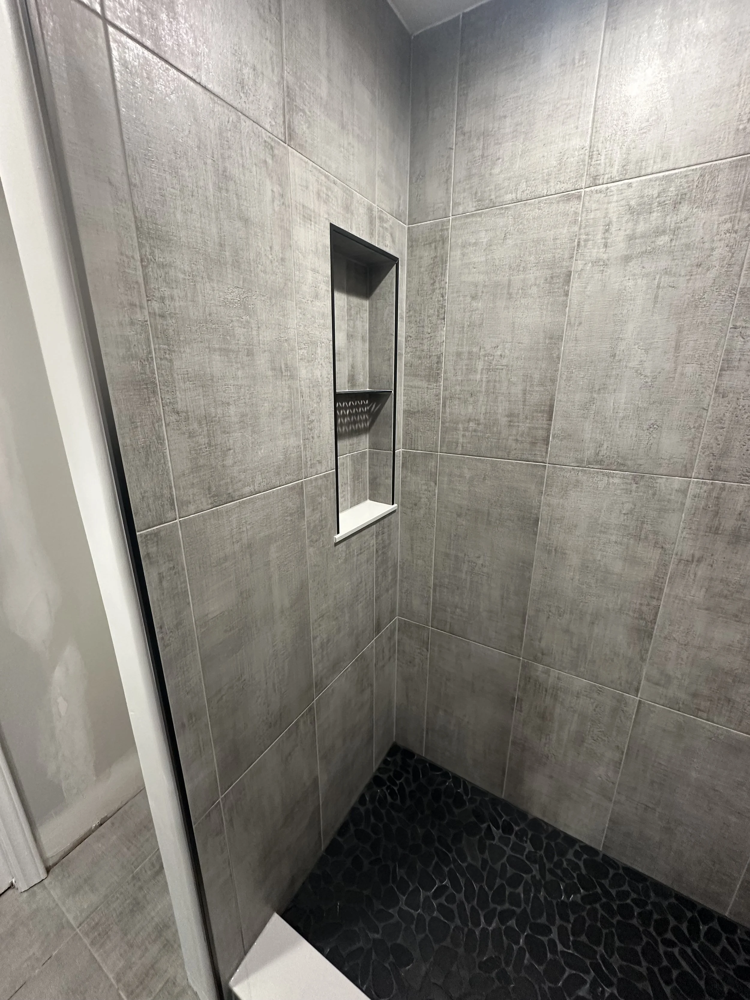 Inside a modern shower with gray tiled walls, a built-in rectangular niche with a small shelf, and a black pebble stone floor.