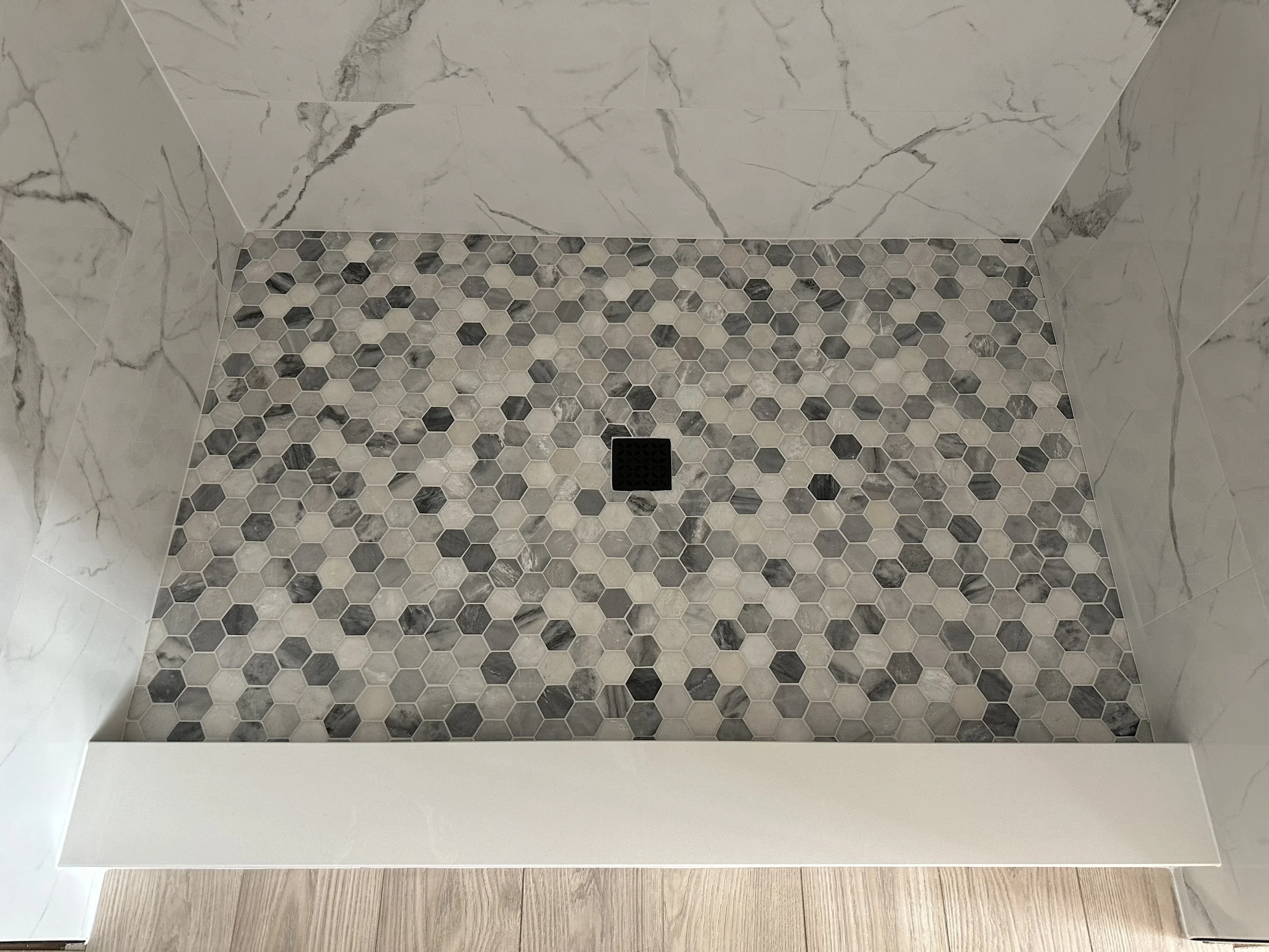Shower floor with small hexagonal tiles in shades of gray and white, black drain cover, surrounded by marble wall tiles.