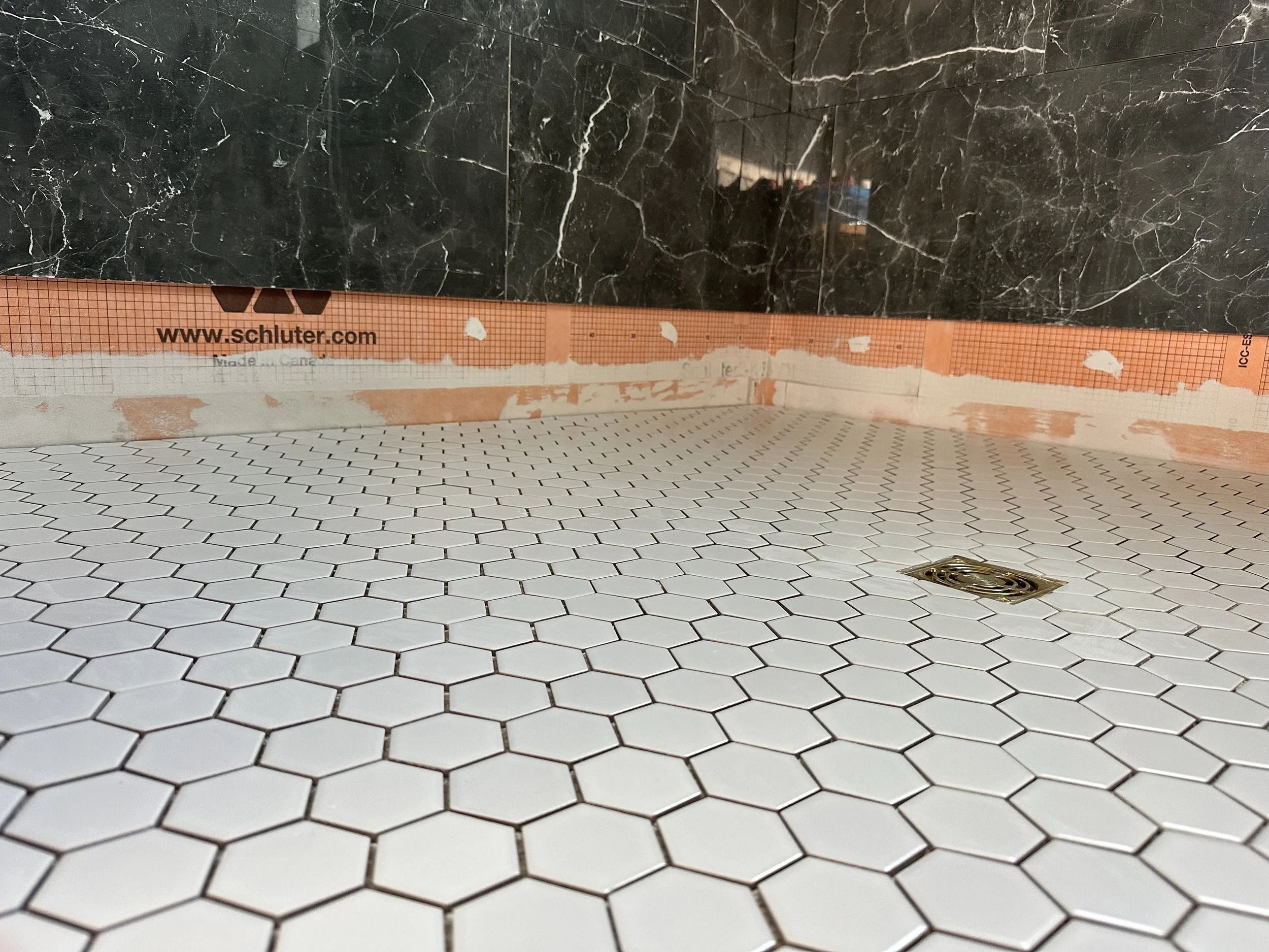 Bathroom floor with white hexagonal tiles, a floor drain, black marble wall, and a cardboard bubble wrap underlayment with site information.
