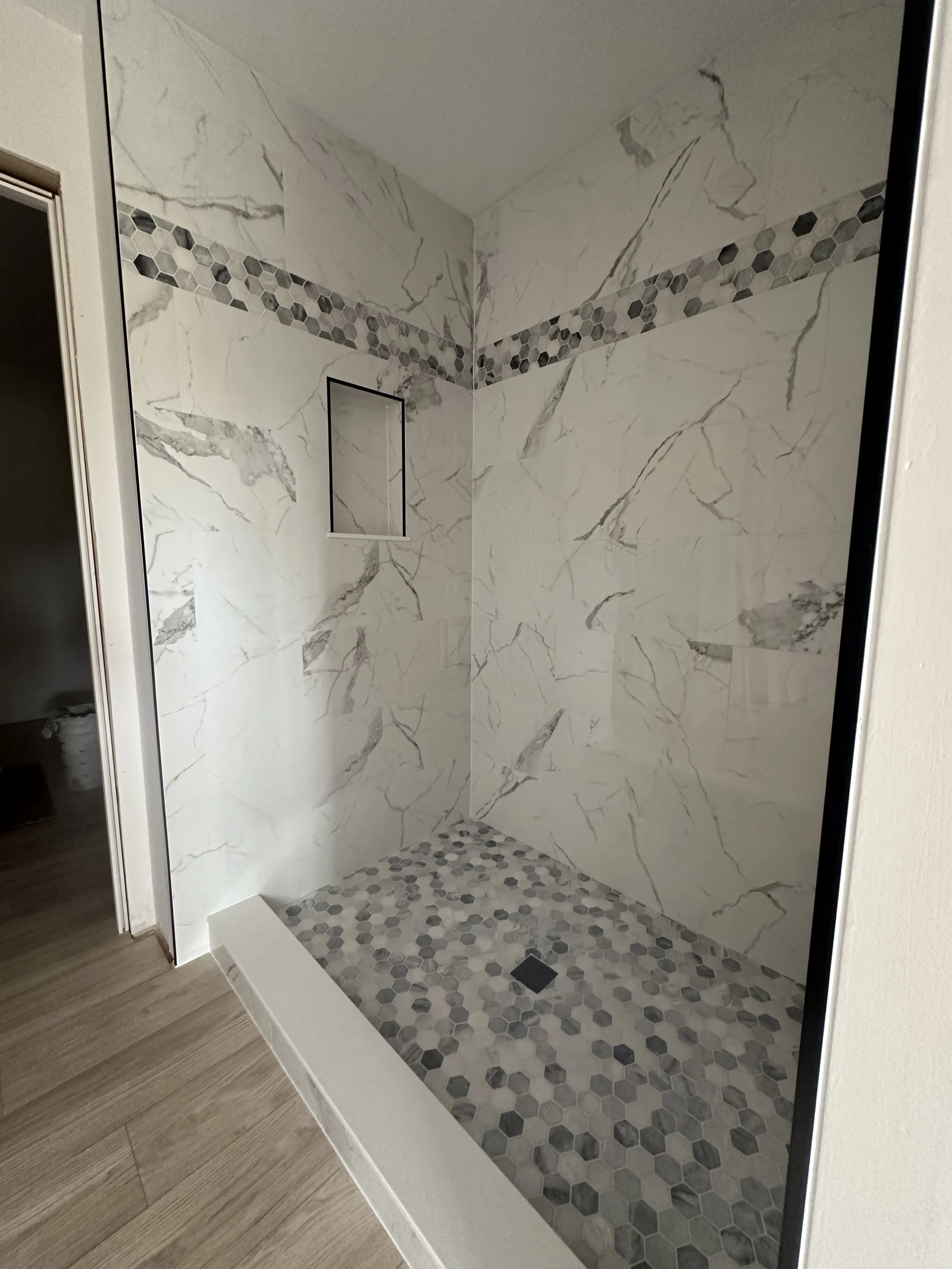 Empty marble shower with a decorative border, niche, and hexagonal tile floor, adjacent to wood flooring.