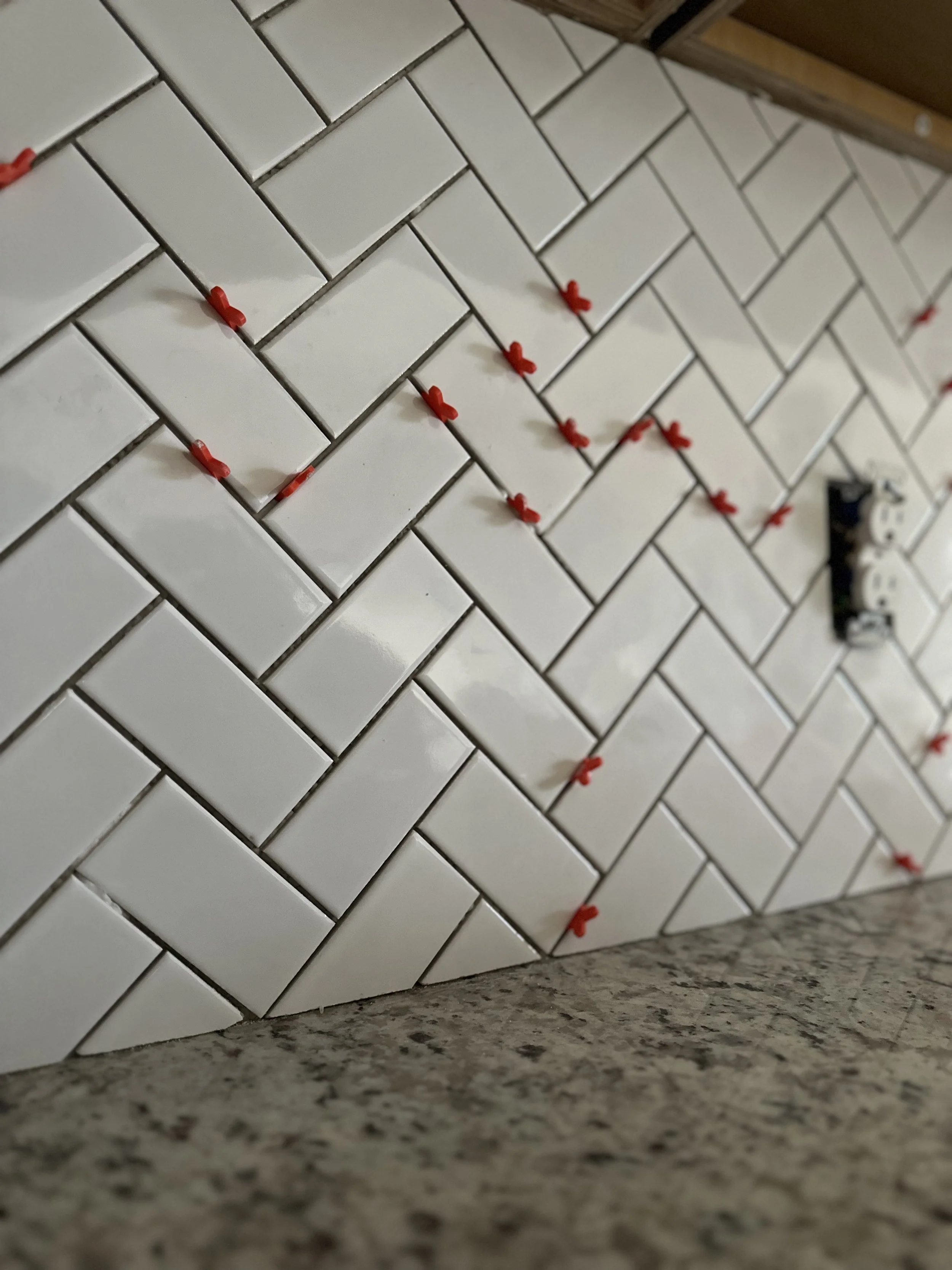 Close-up of a tiled wall with white rectangular tiles arranged in a herringbone pattern, with small red spacers inserted between the tiles for grouting. Part of a granite countertop is visible at the bottom.