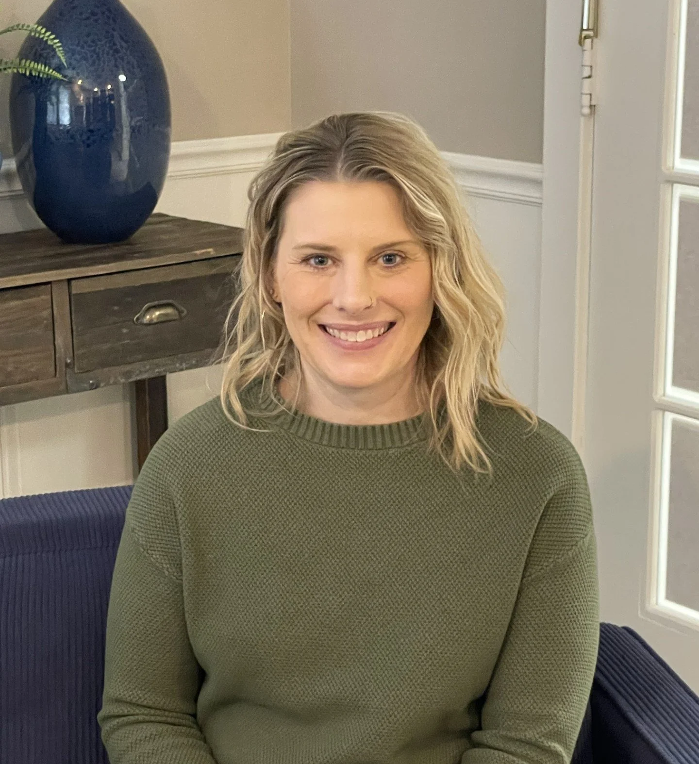 A woman with blonde wavy hair smiling and wearing a green sweater, sitting on a blue chair in a room with neutral decor and a wooden sideboard with a blue vase on top.