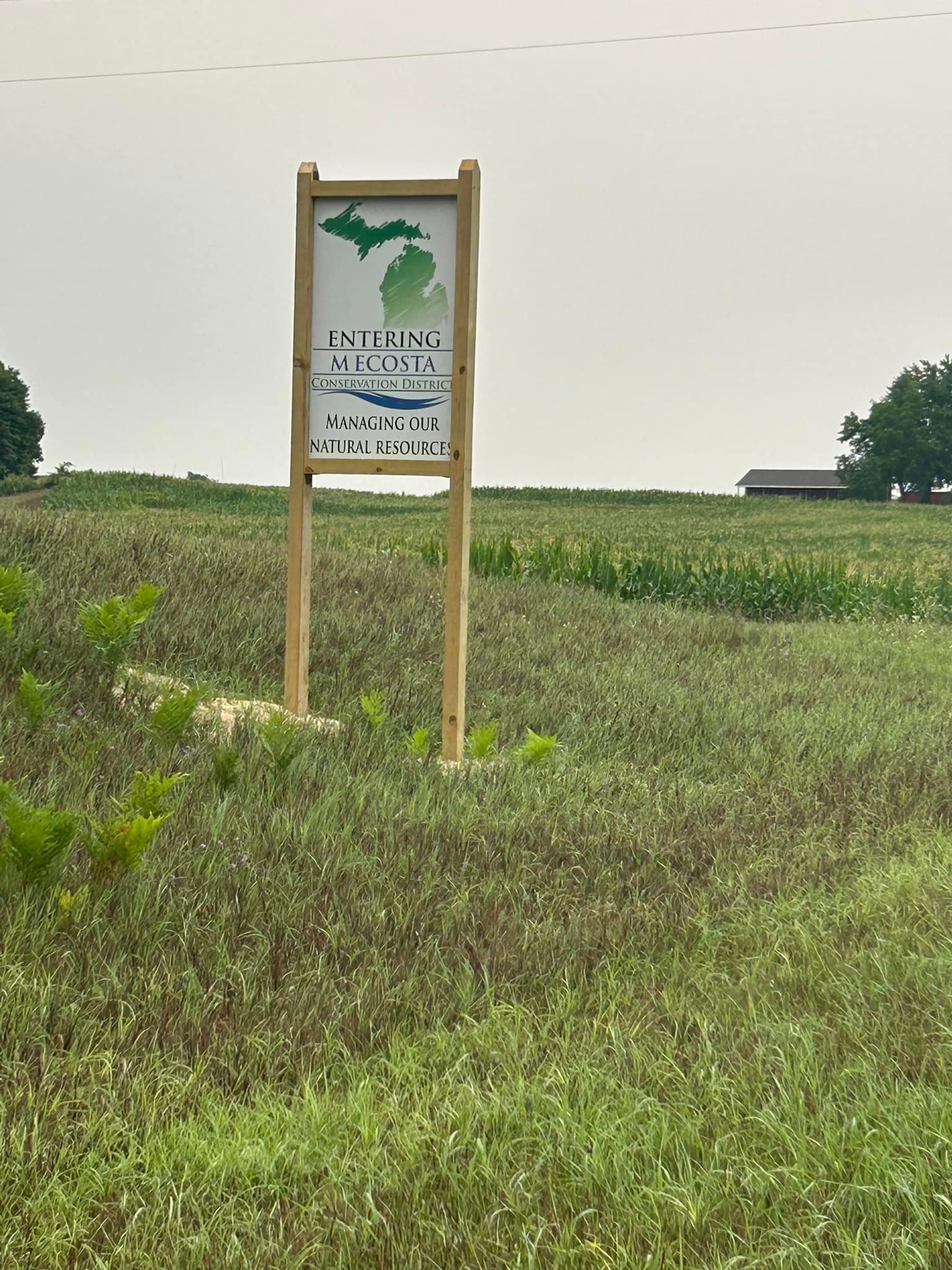 A sign in a grassy field reads: "Entering Mecosta Conservation District, Managing Our Natural Resources," with a map outline in the background. The sky is overcast, and a tree and barn are visible in the distance.