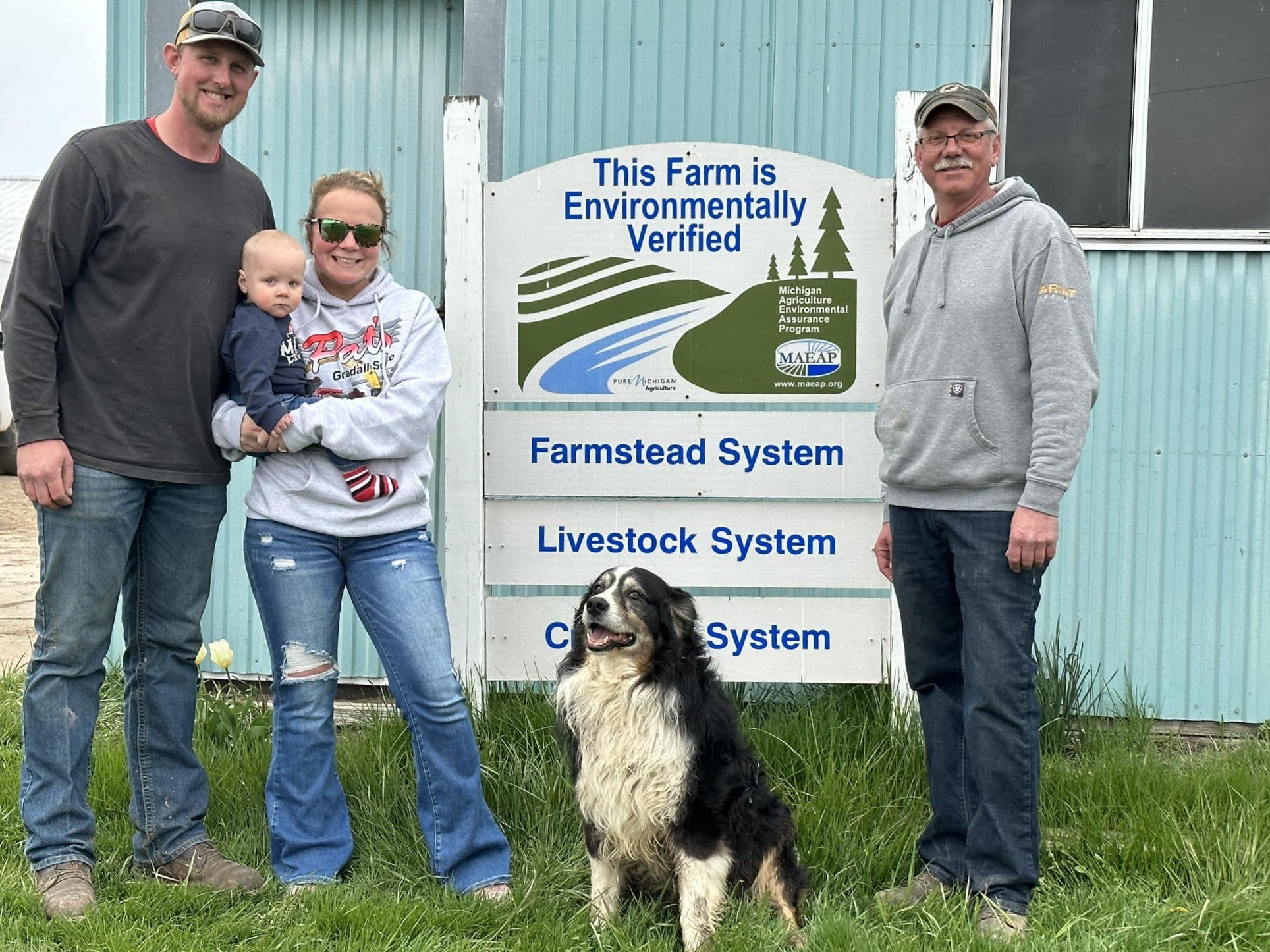 A family standing in front of a sign for a farm certified as environmentally verified by the Michigan Agriculture Environmental Assurance Program. A dog is also present. The adults and baby are smiling, and the dog is sitting on the grass. The backgr