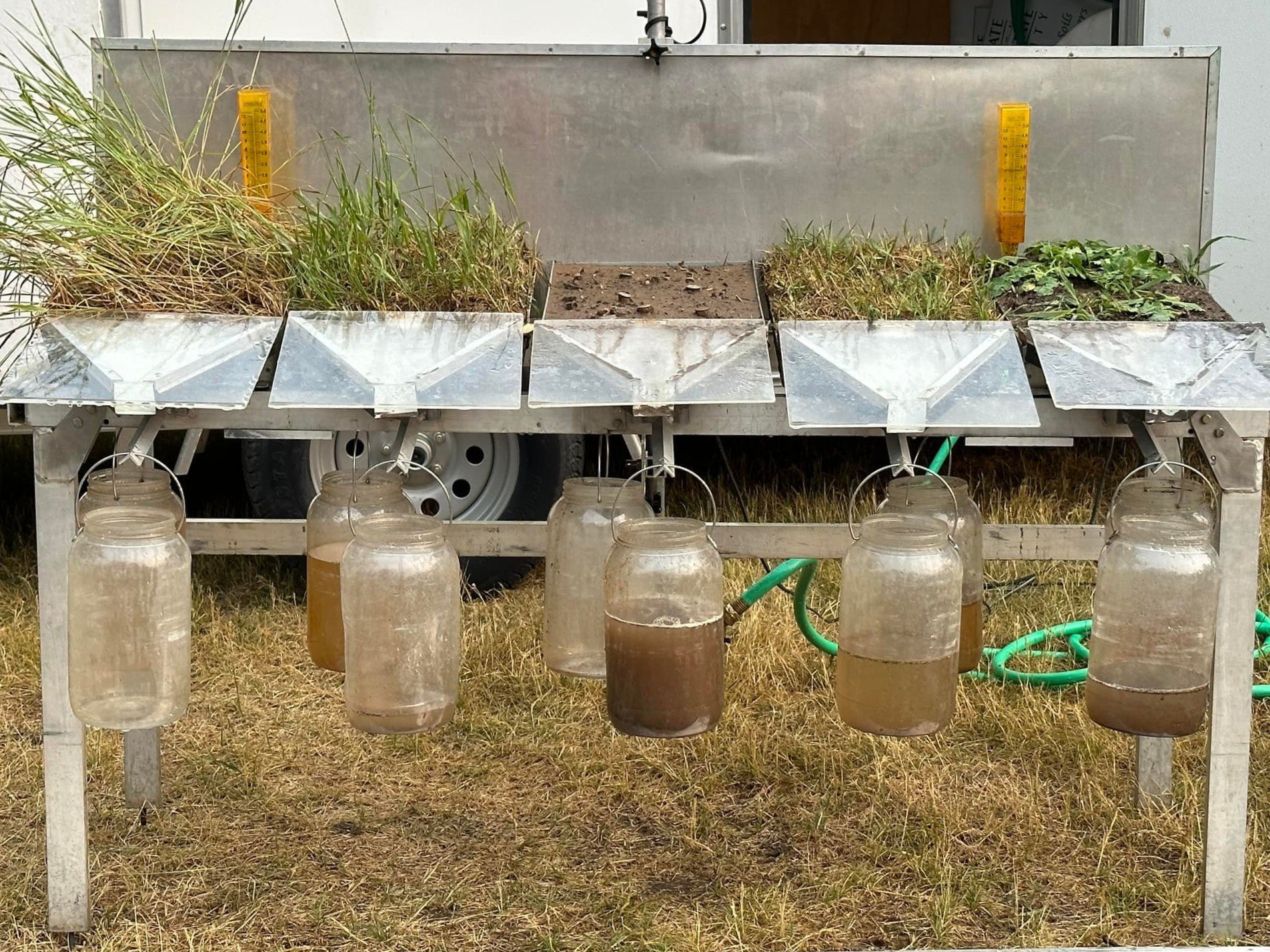Soil erosion demonstration setup with jars collecting runoff water from different soil types and vegetation, showing varying water turbidity levels.