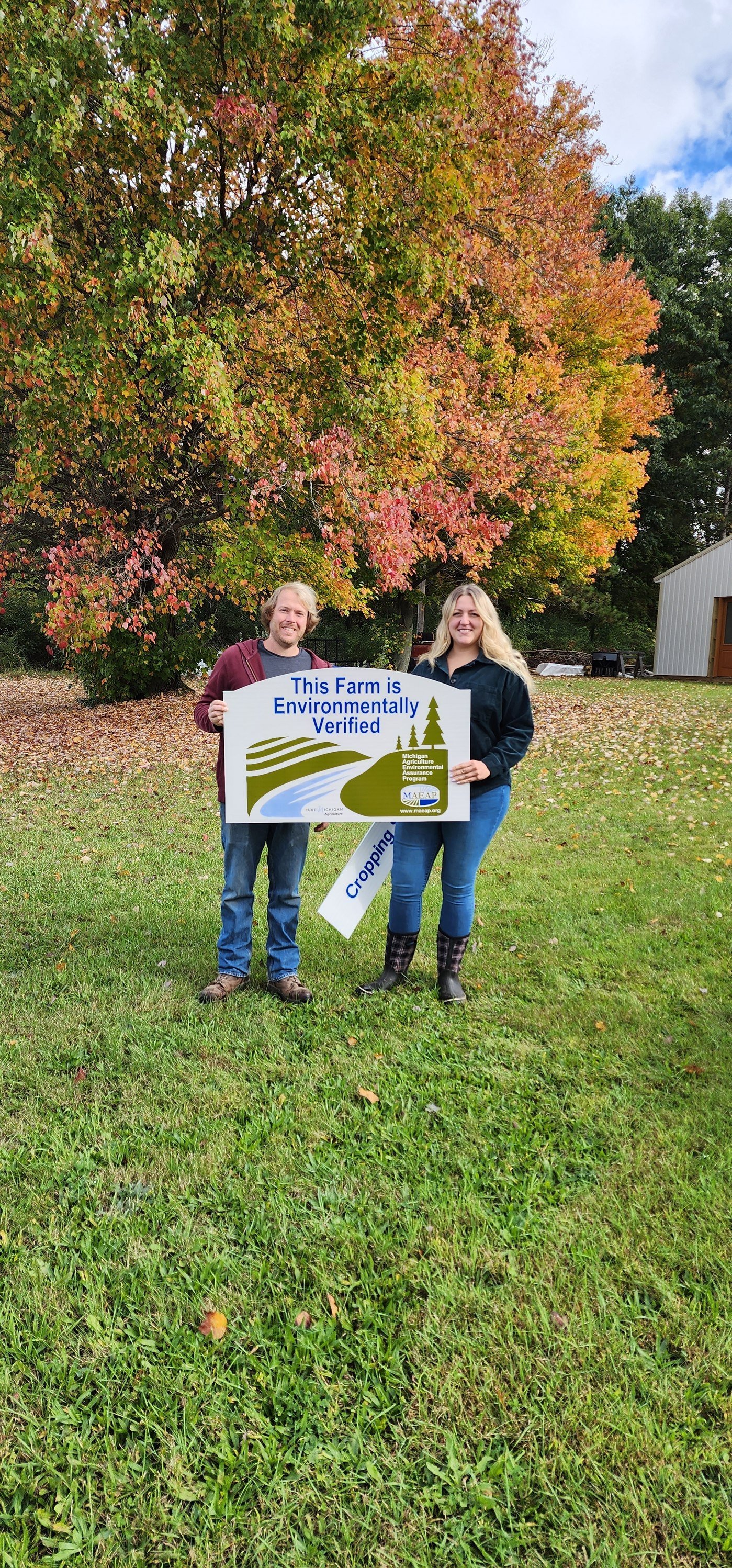 Two people standing on a grassy area in front of a tree with autumn leaves, holding a sign that says 'This Farm is Environmentally Verified' by the Michigan Agriculture Environmental Assurance Program (MAEAP).